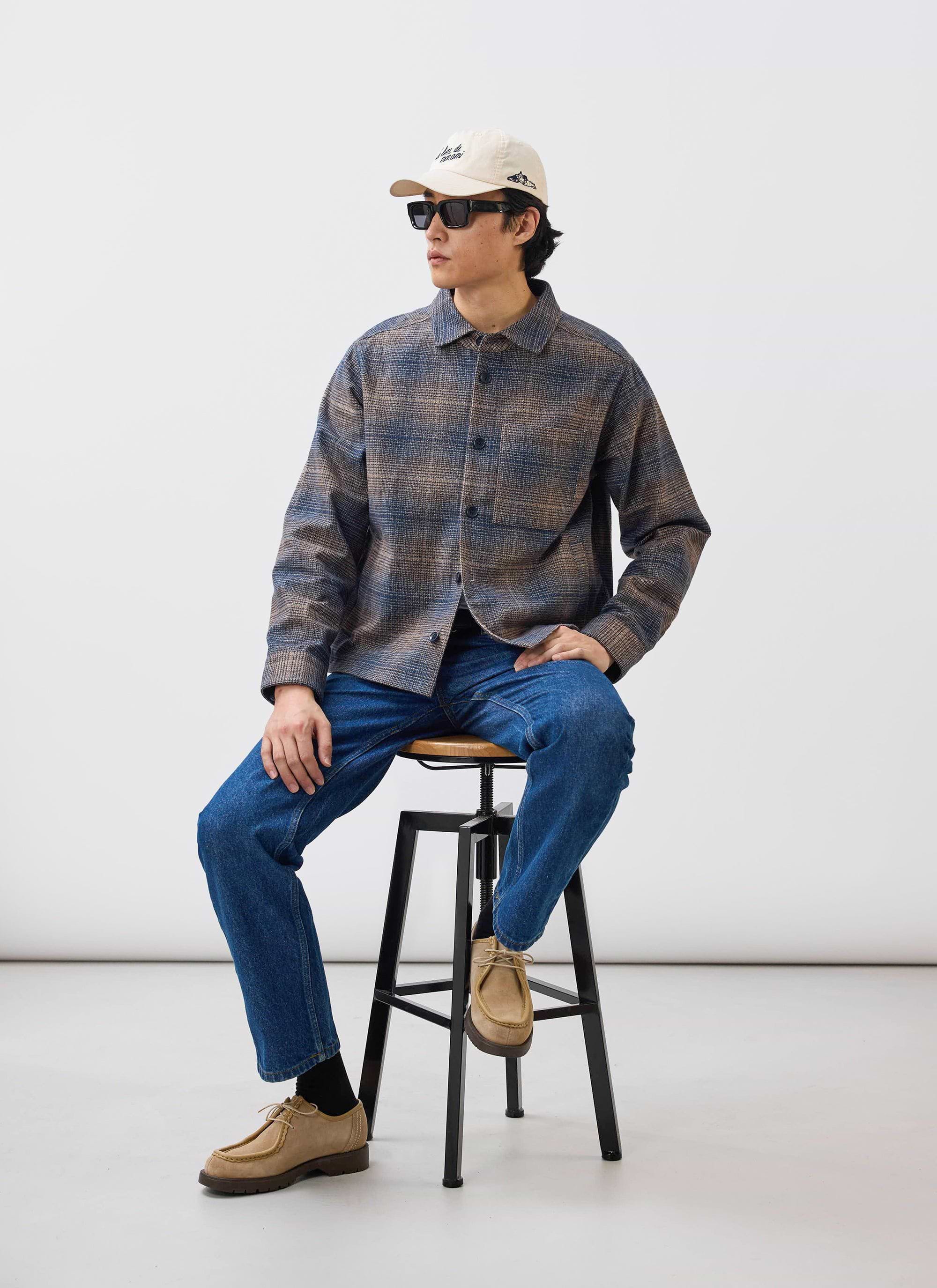 A model wears the Percival Luton Check Overshirt in brown, styled with blue jeans, a cap and sunglasses while sat on a stool.