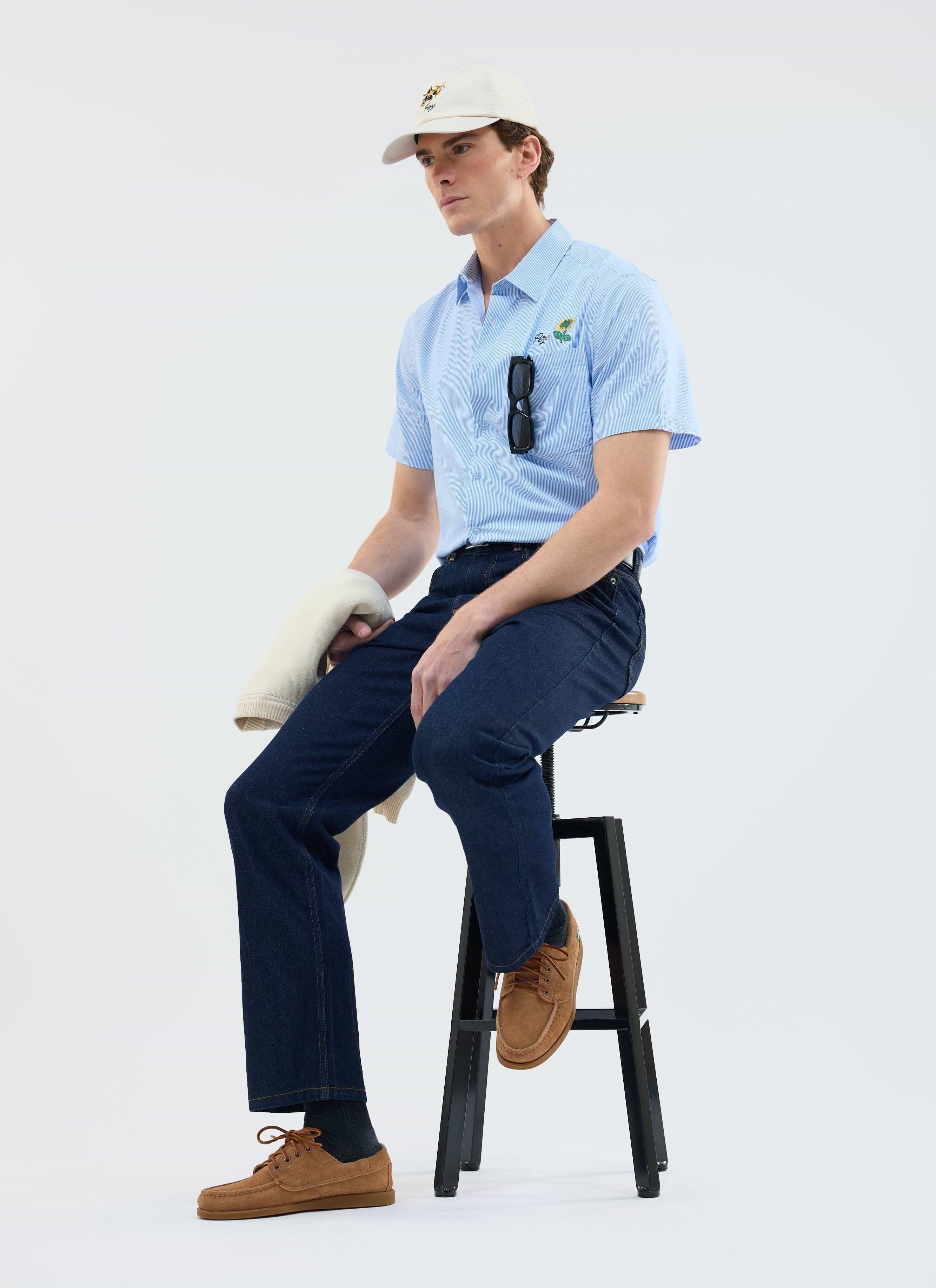 A model wears the Percival Sunflower Stripe Shirt in blue with dark jeans, seated on a stool in a studio.