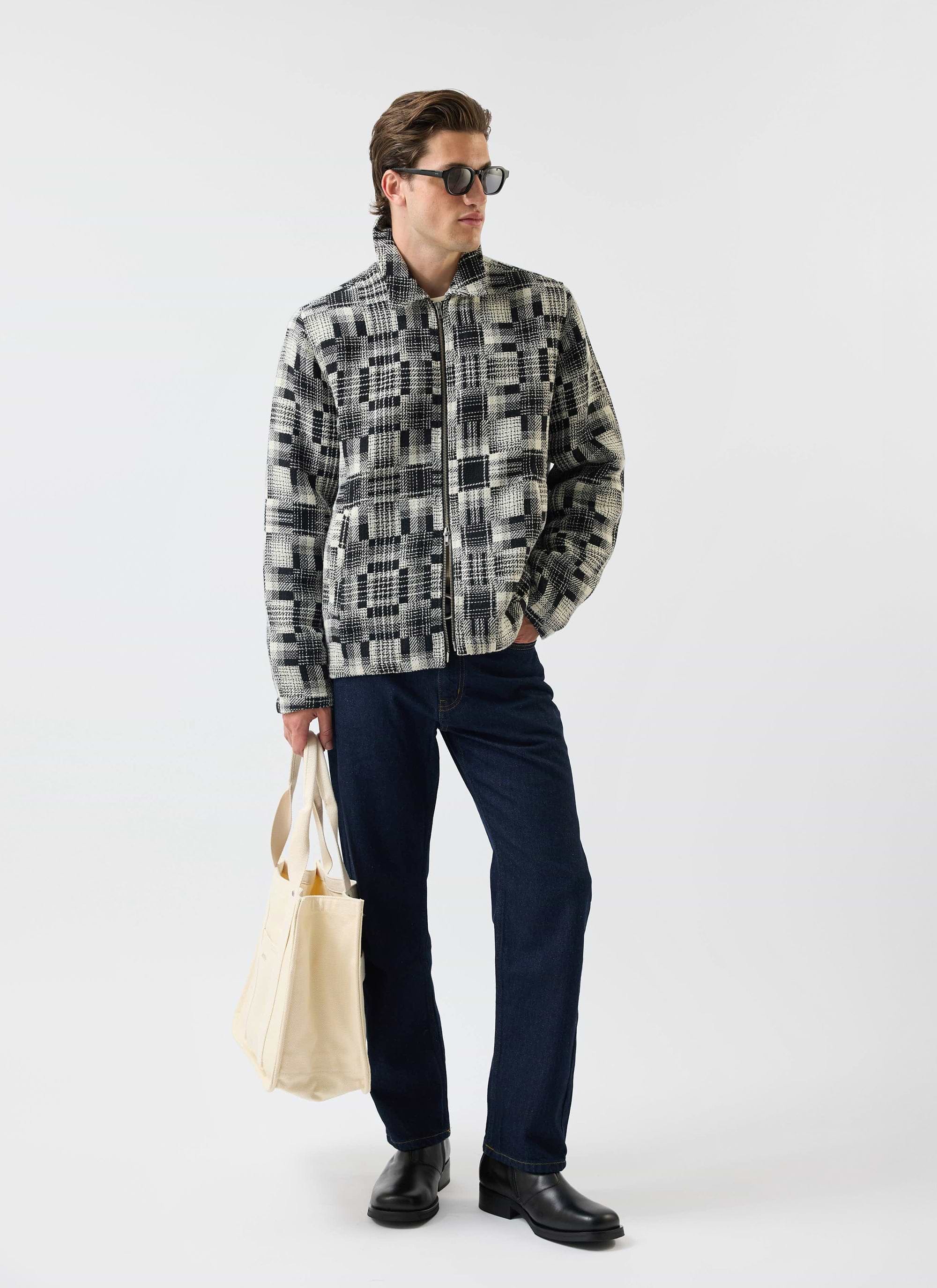A model wears the Percival Glossary Patchwork Jacket in grey twill, styled with dark denim, sunglasses, and a tote bag.