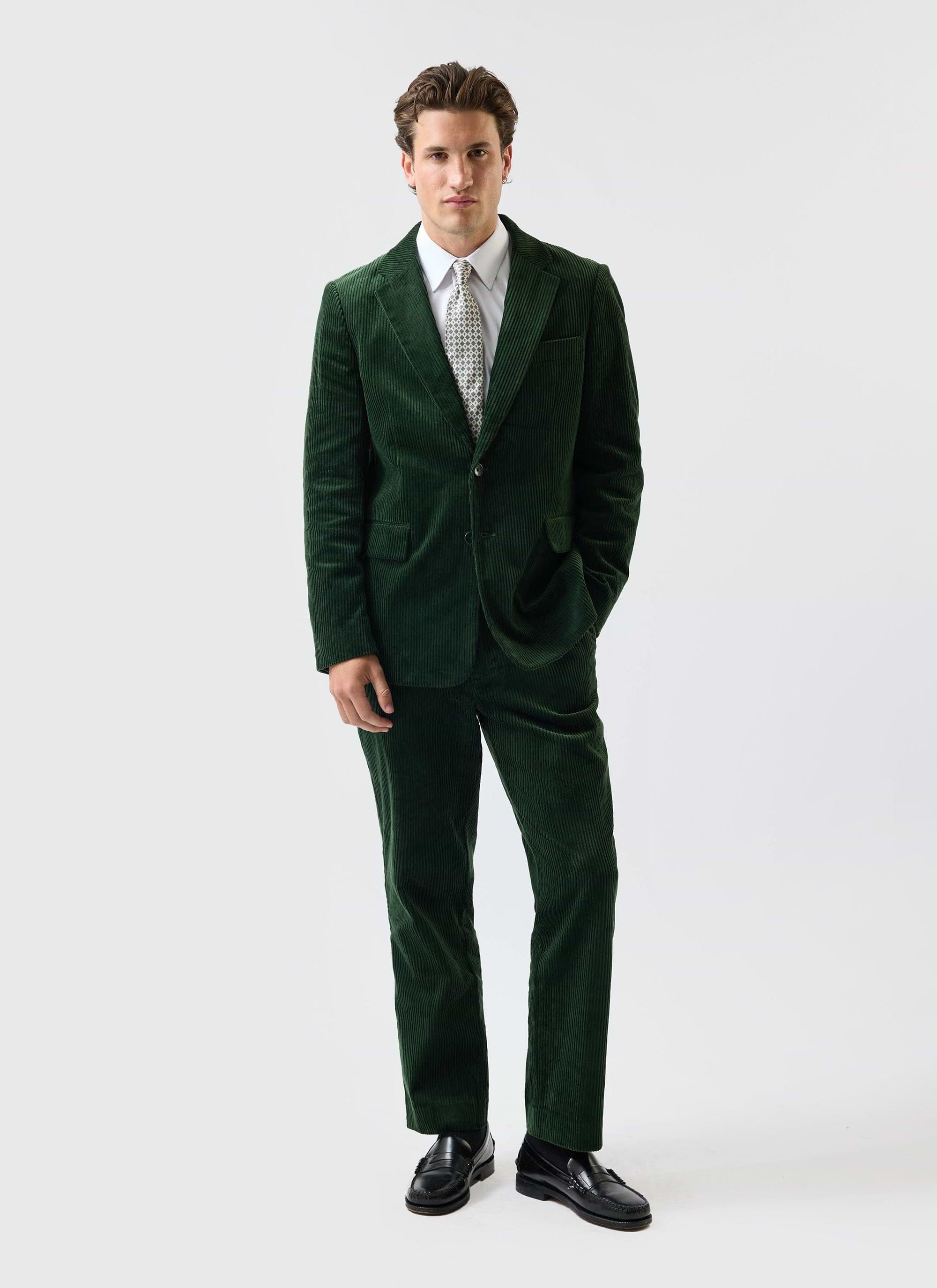 A model wears the Percival Chunky Cord suit in forest green with a white shirt, patterned tie, and black loafers.