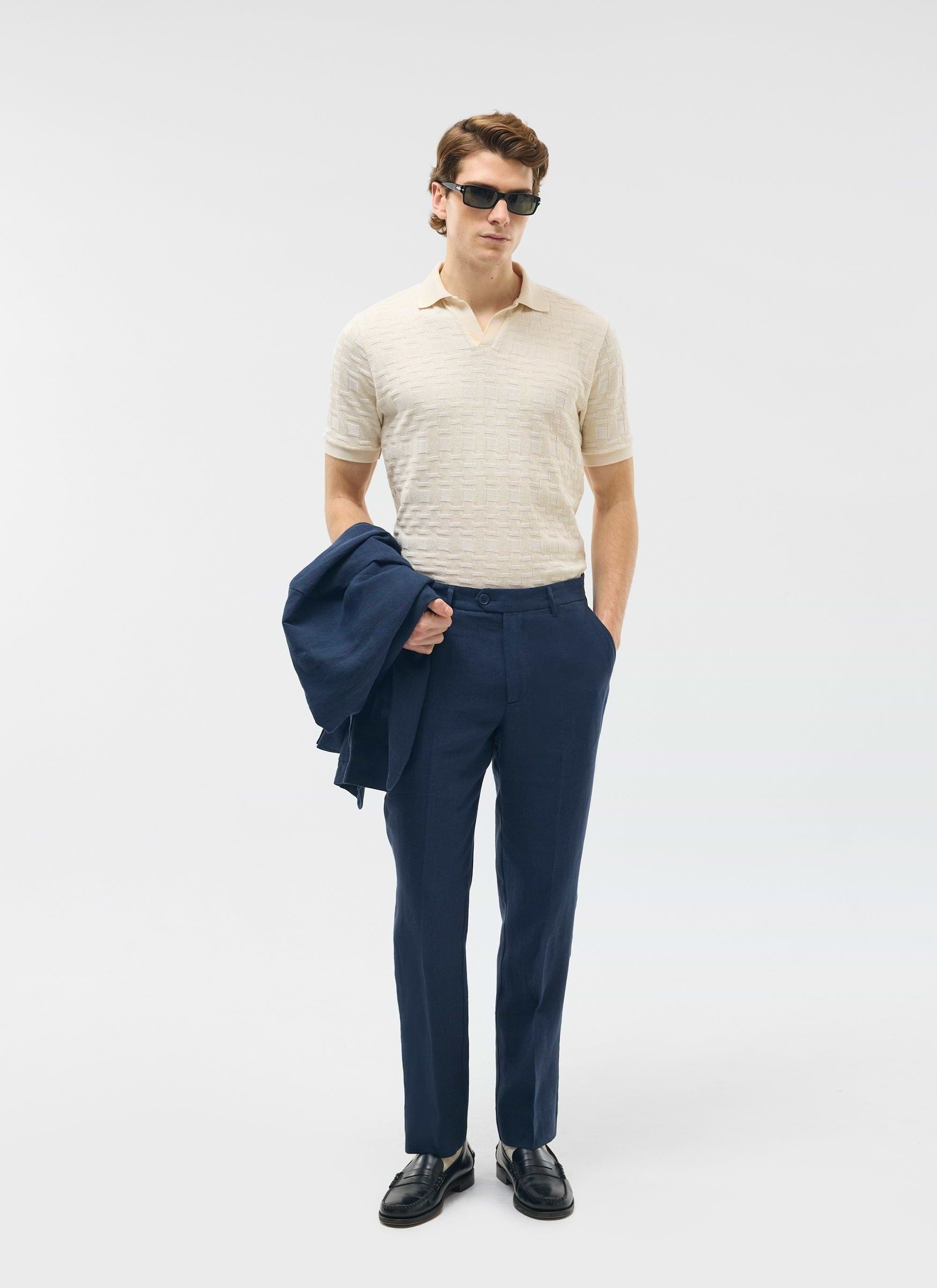 A model wears Percival's navy tailored linen trousers with a cream polo shirt, holding the matching navy jacket.