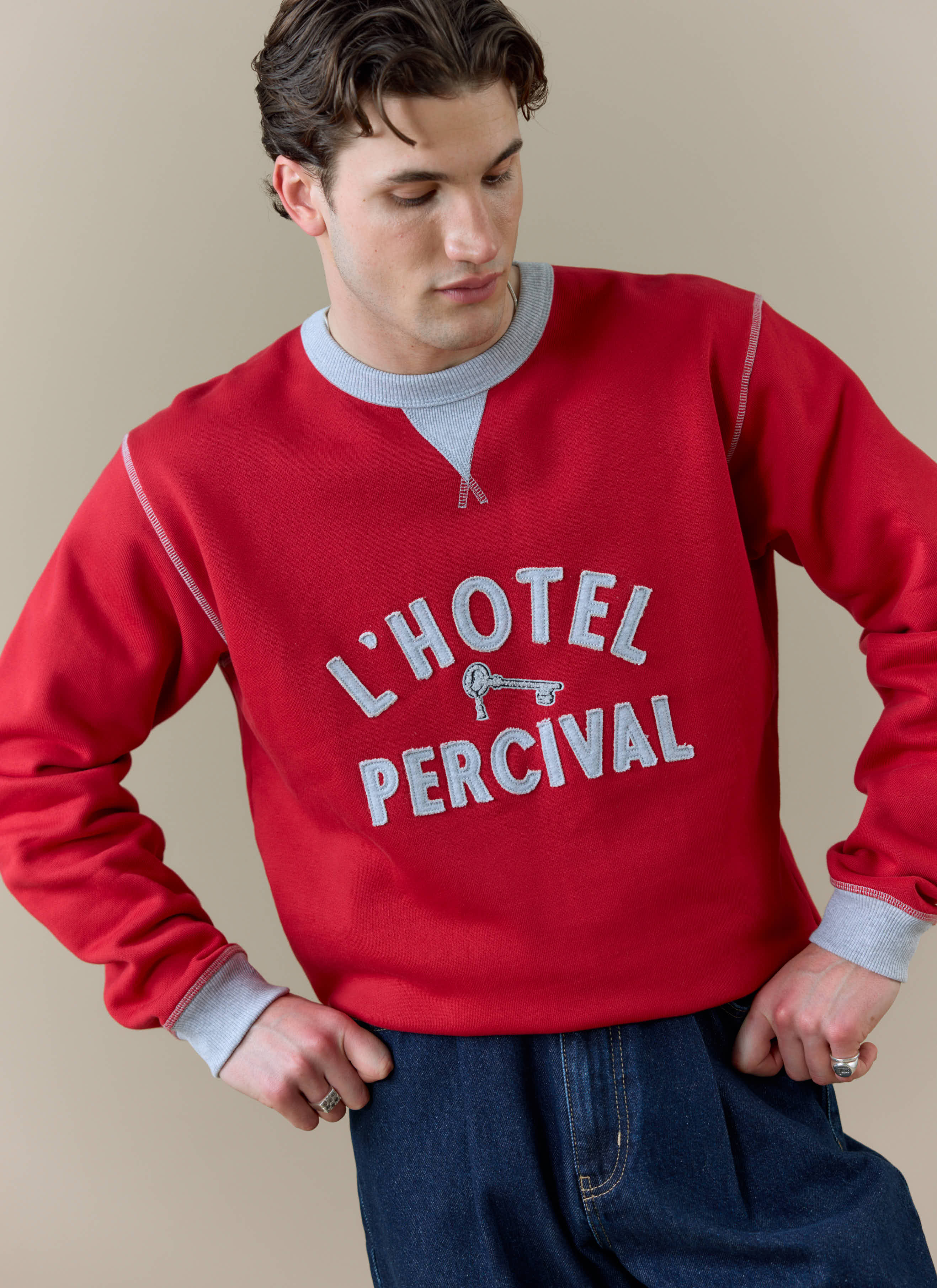 Close-up of a model wearing the red Percival Ronald Sweatshirt, featuring the grey "L'HOTEL PERCIVAL" applique design.