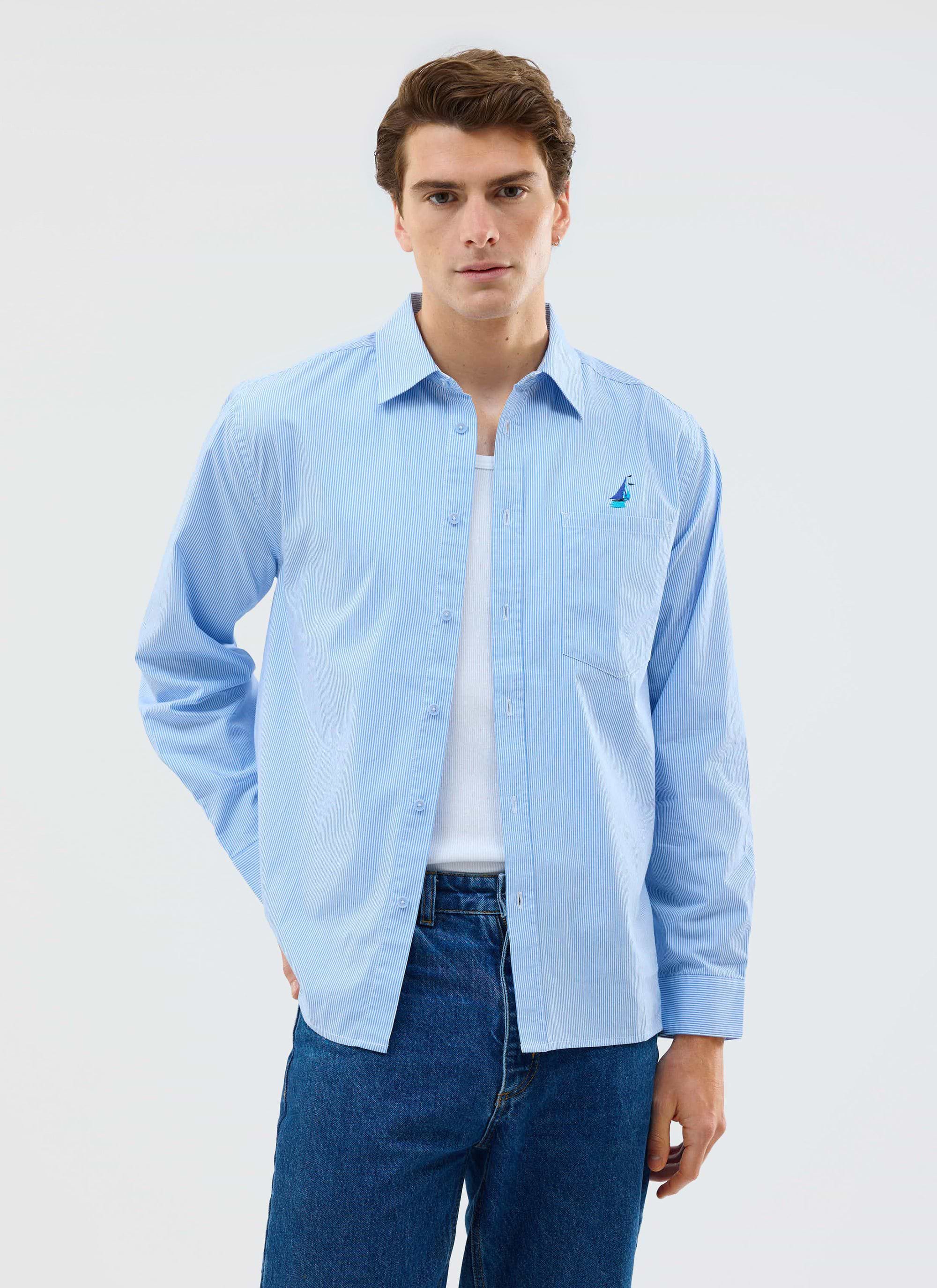 A man models the Percival blue Yacht Stripe Oversized Shirt, open over a white vest, styled with dark blue jeans.