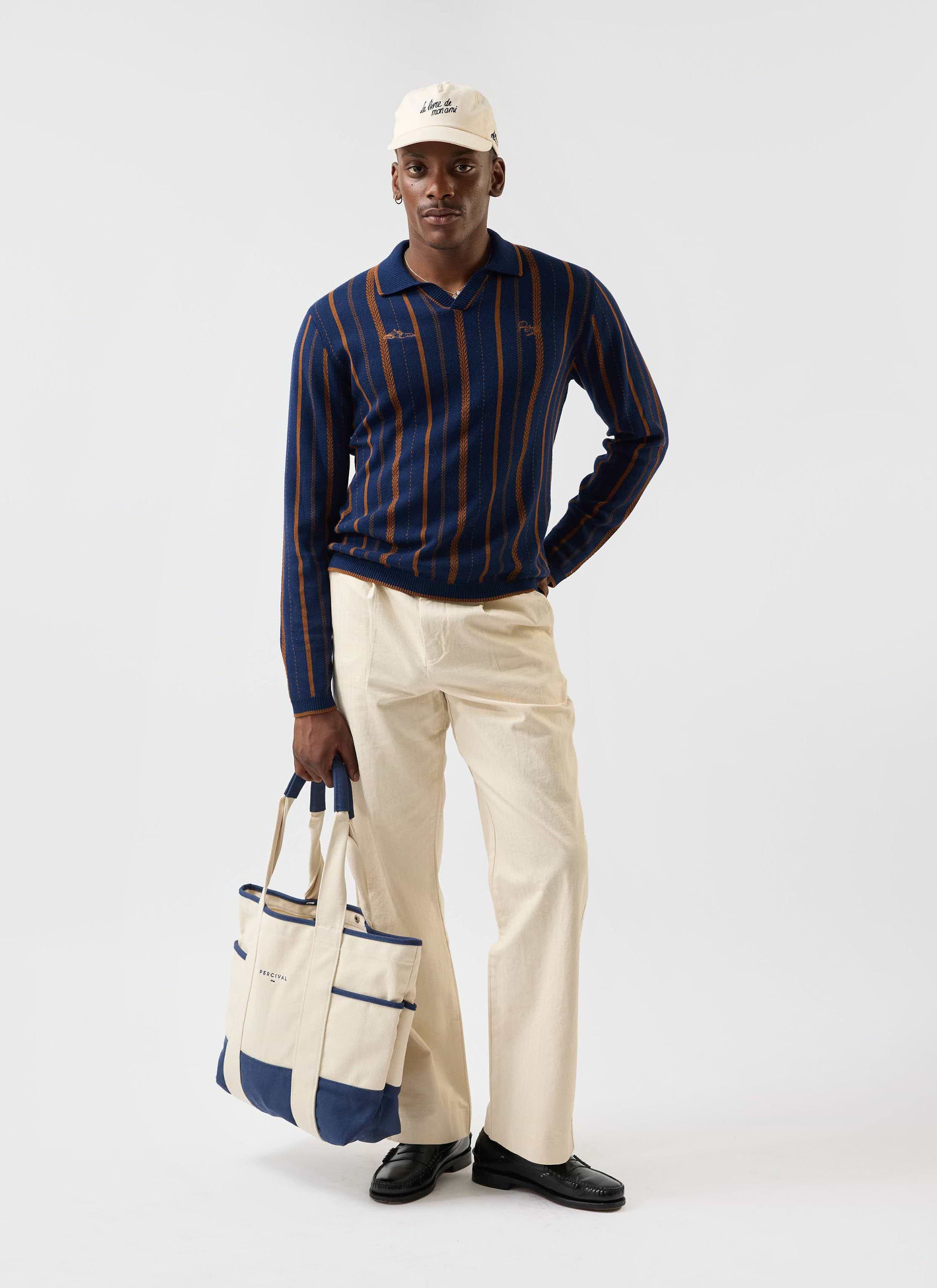 A model wears the navy Percival Citation Knitted Football Shirt, styled with cream trousers, a cap, and a canvas tote bag.