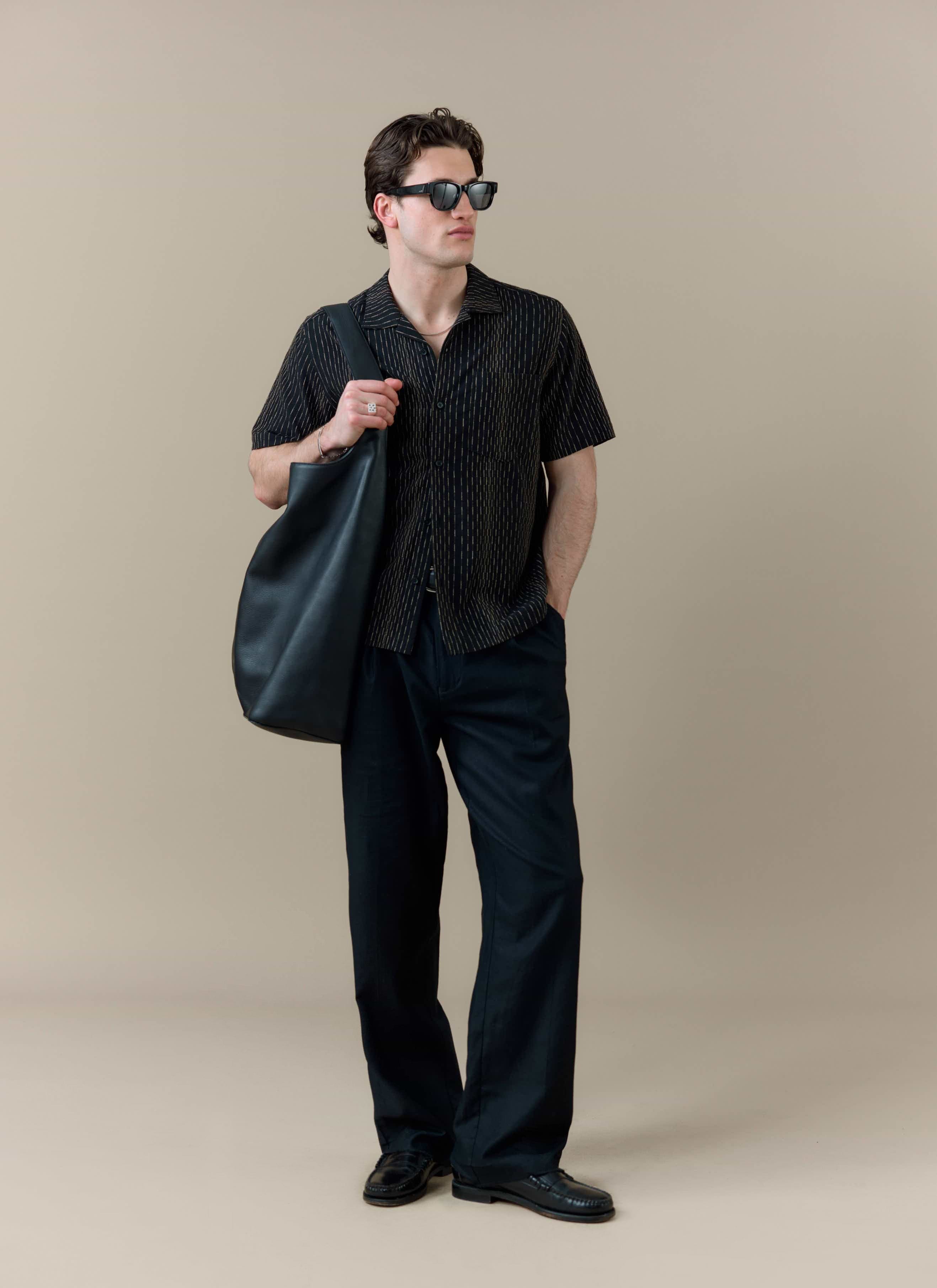 A model wears the black cotton Percival Leland Cuban Shirt with vertical stripes, black trousers and sunglasses.