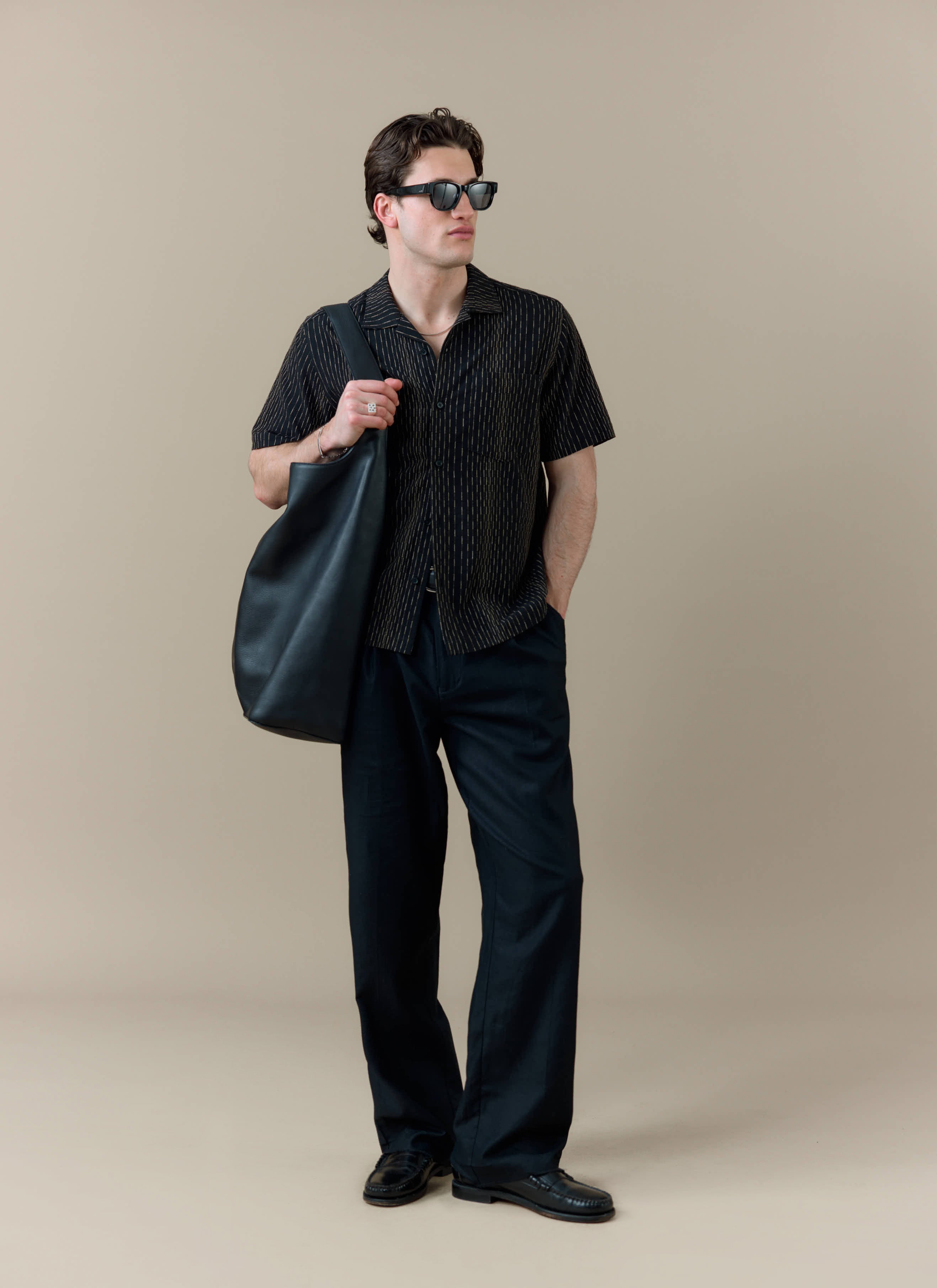 A model wears the black cotton Percival Leland Cuban Shirt with vertical stripes, black trousers and sunglasses.