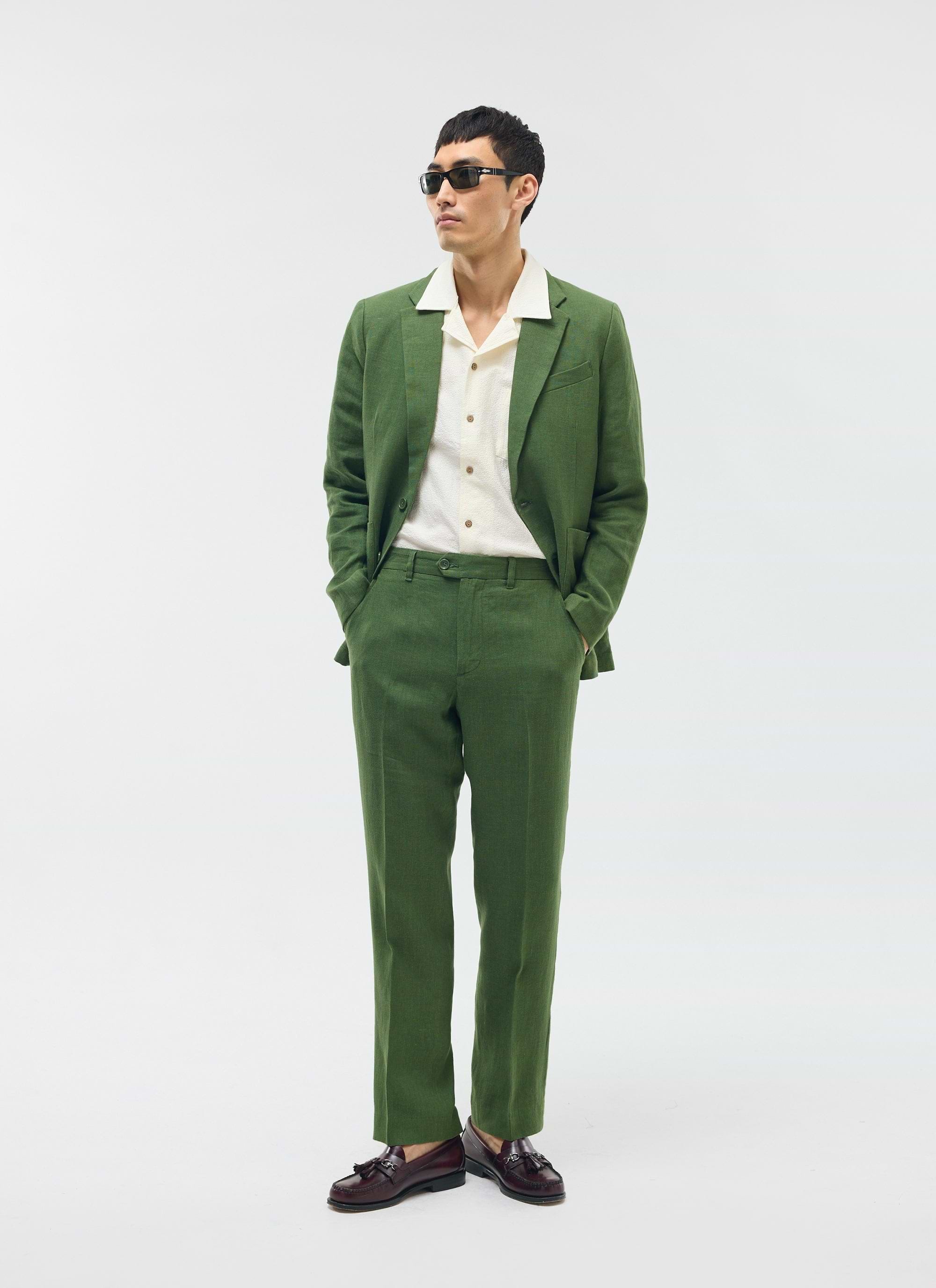 A model wears a Percival forest green tailored linen suit with a cream shirt and sunglasses against a white background.