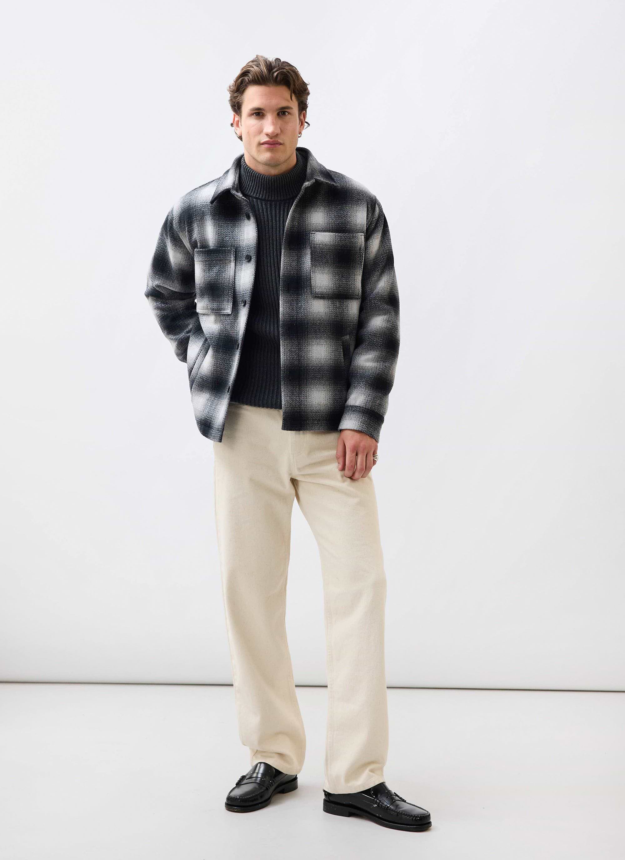 A man models the Percival Check Overshirt in grey, styled with a dark grey turtleneck, cream trousers and black loafers.