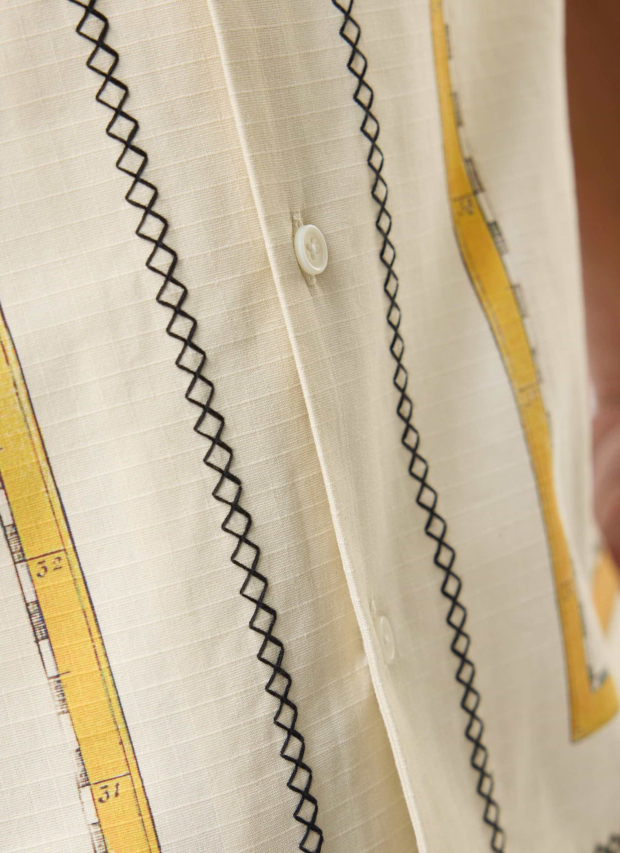 A close-up of the Percival Ex Libris Cuban Shirt, showing the ecru cotton ripstop, black cross-stitch embroidery and artwork.