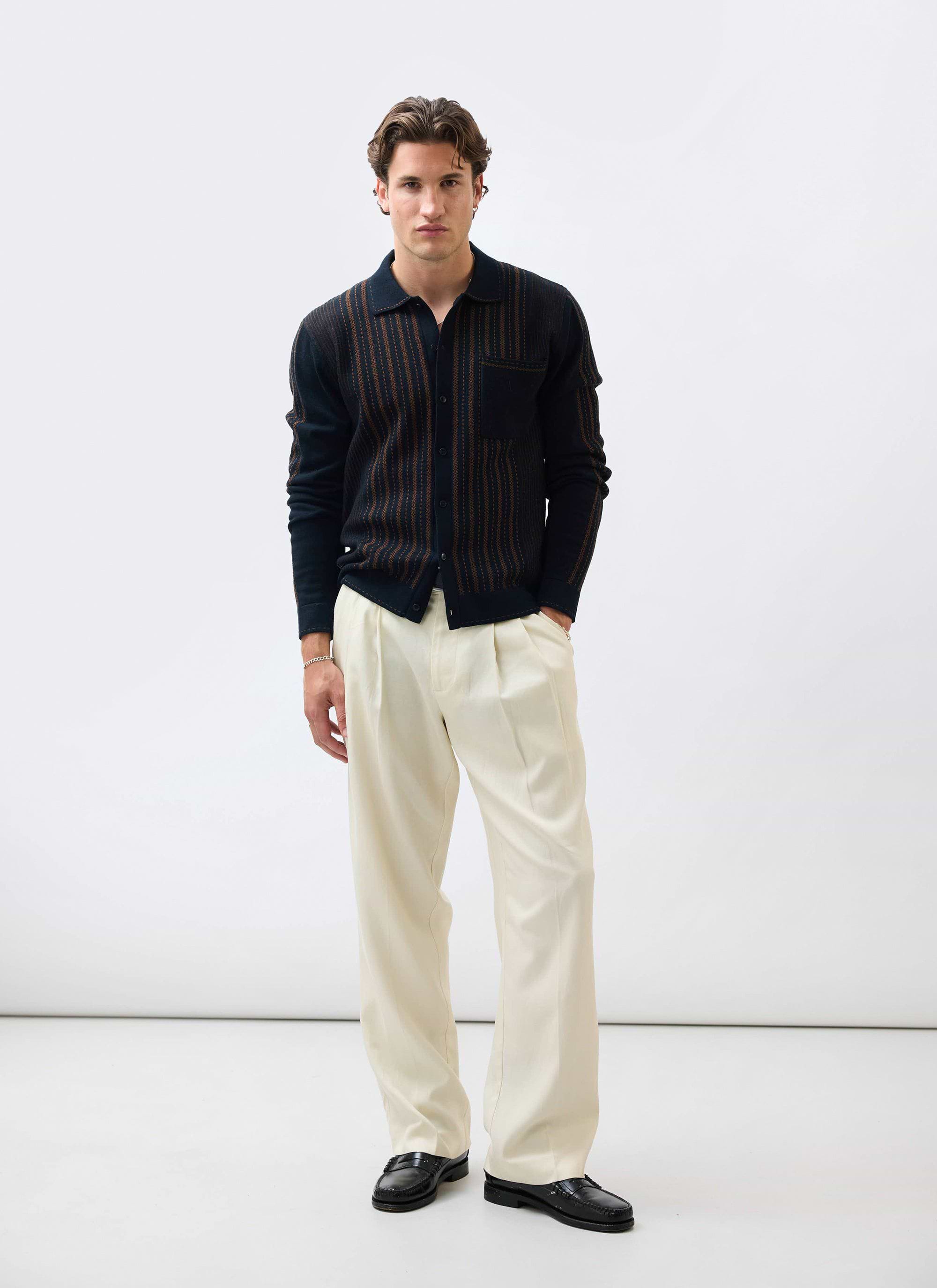 A model wears the Percival Appendix Long Sleeve Knitted Shirt in black and brown with cream trousers and black loafers.