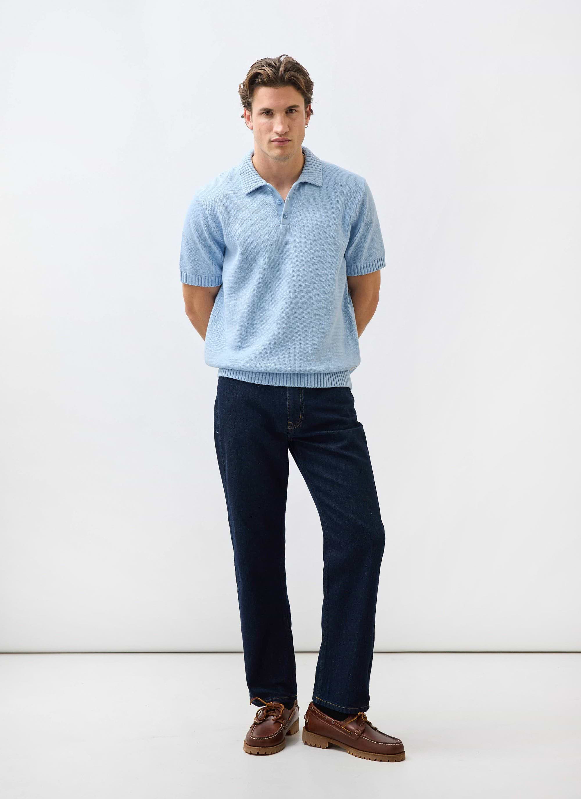 A man models the Percival Verso Short Sleeve Knitted Shirt in Heather Blue with dark jeans and brown boat shoes.
