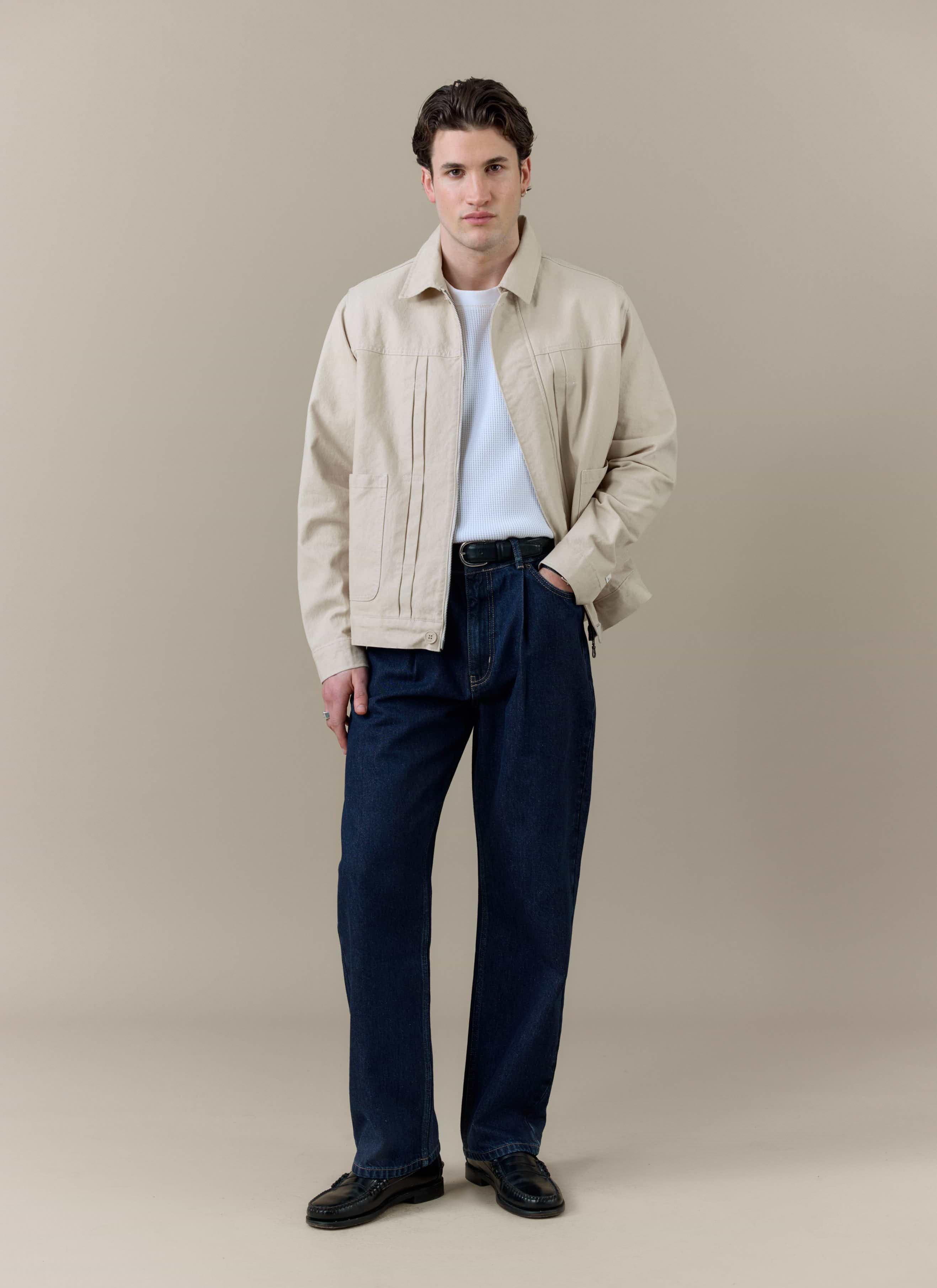A model wearing the Percival Ernst Jacket in cream cotton, featuring front pleats, styled with dark denim jeans.