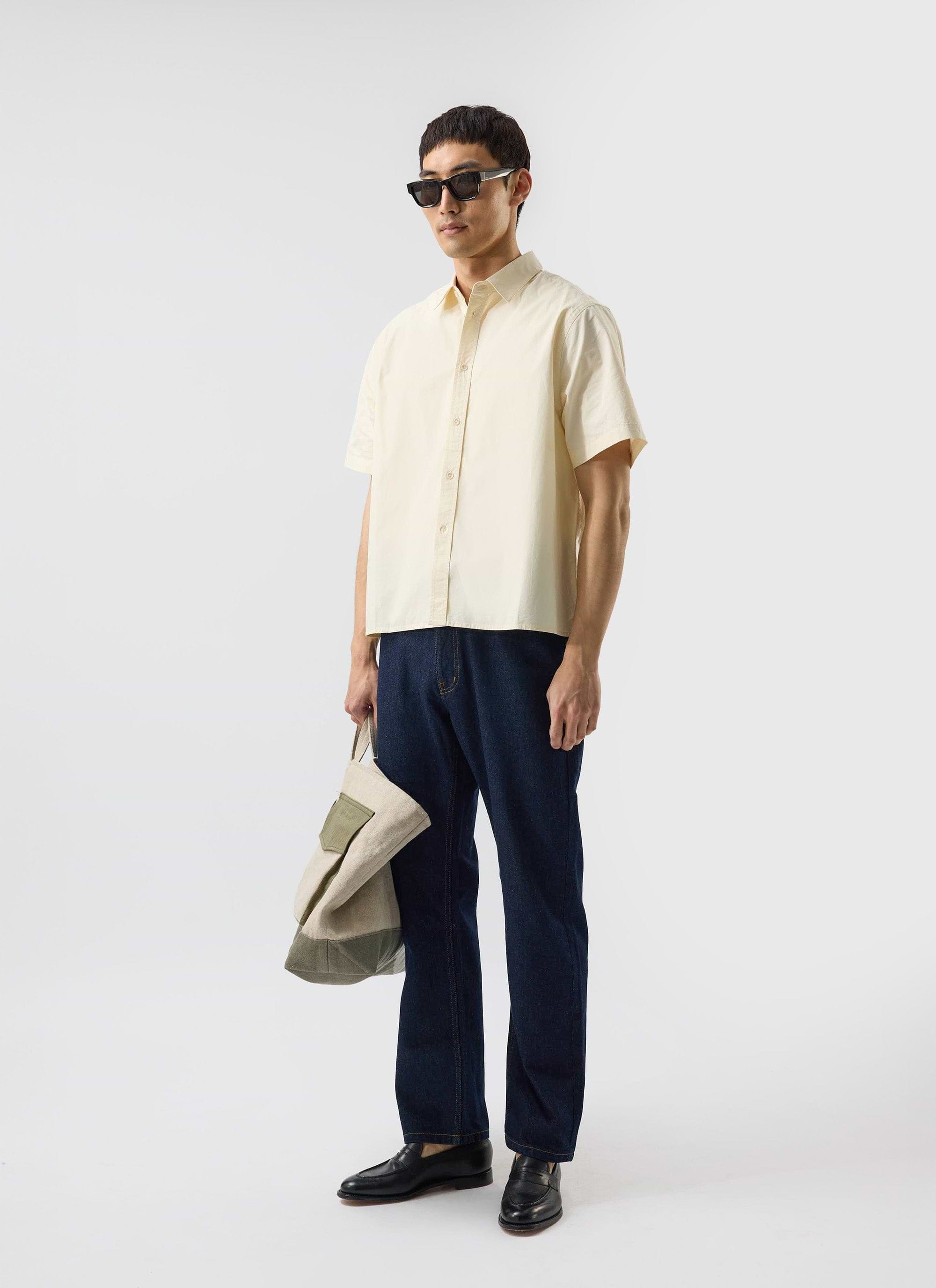 A man models the Percival Boxy Cropped Clerk Shirt in ecru, with dark jeans, black loafers and sunglasses.