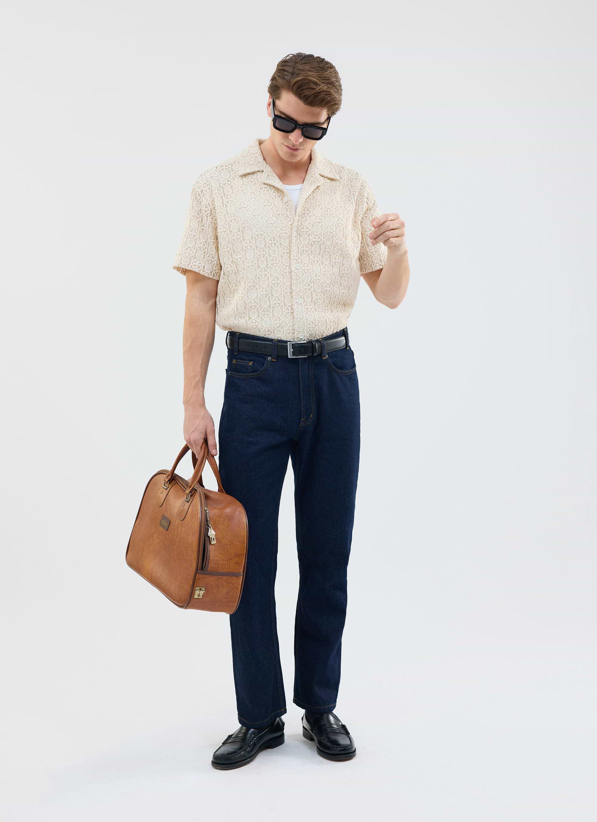 A model wears the Percival Elio Crochet Cuban Shirt in ecru with dark denim, sunglasses, and a leather weekend bag.