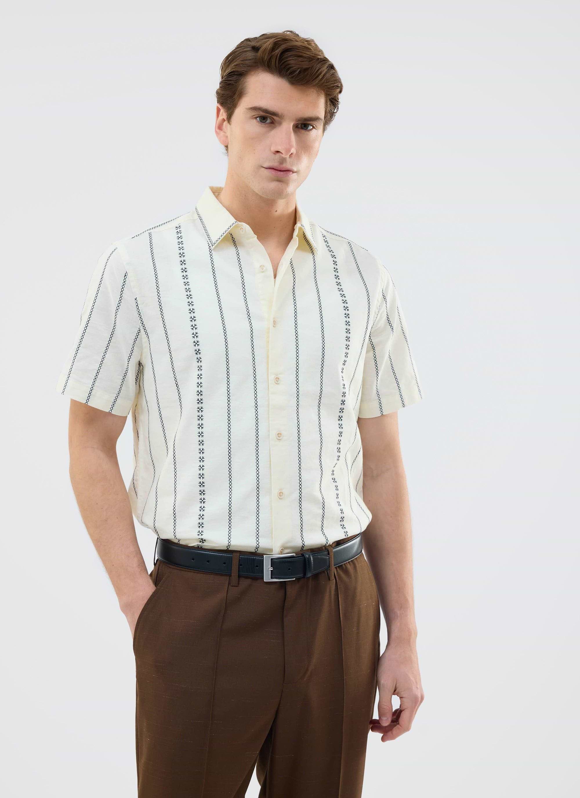 A man models the Percival Cross Stitch Boxy Clerk Shirt in ecru, styled with smart brown trousers.