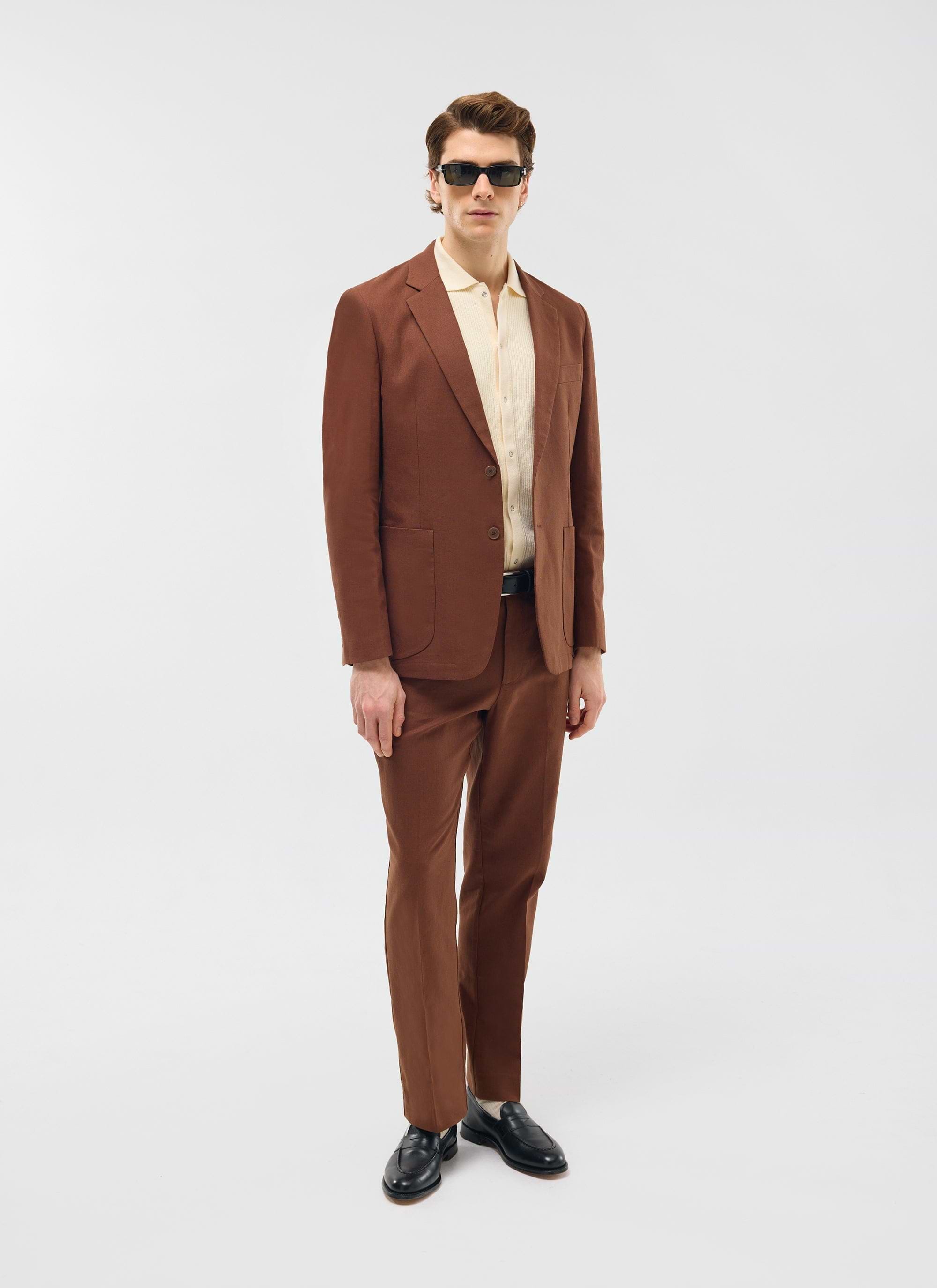 A model wears the Percival Tailored Linen Trousers in brown with the matching blazer, a cream knit shirt and sunglasses.