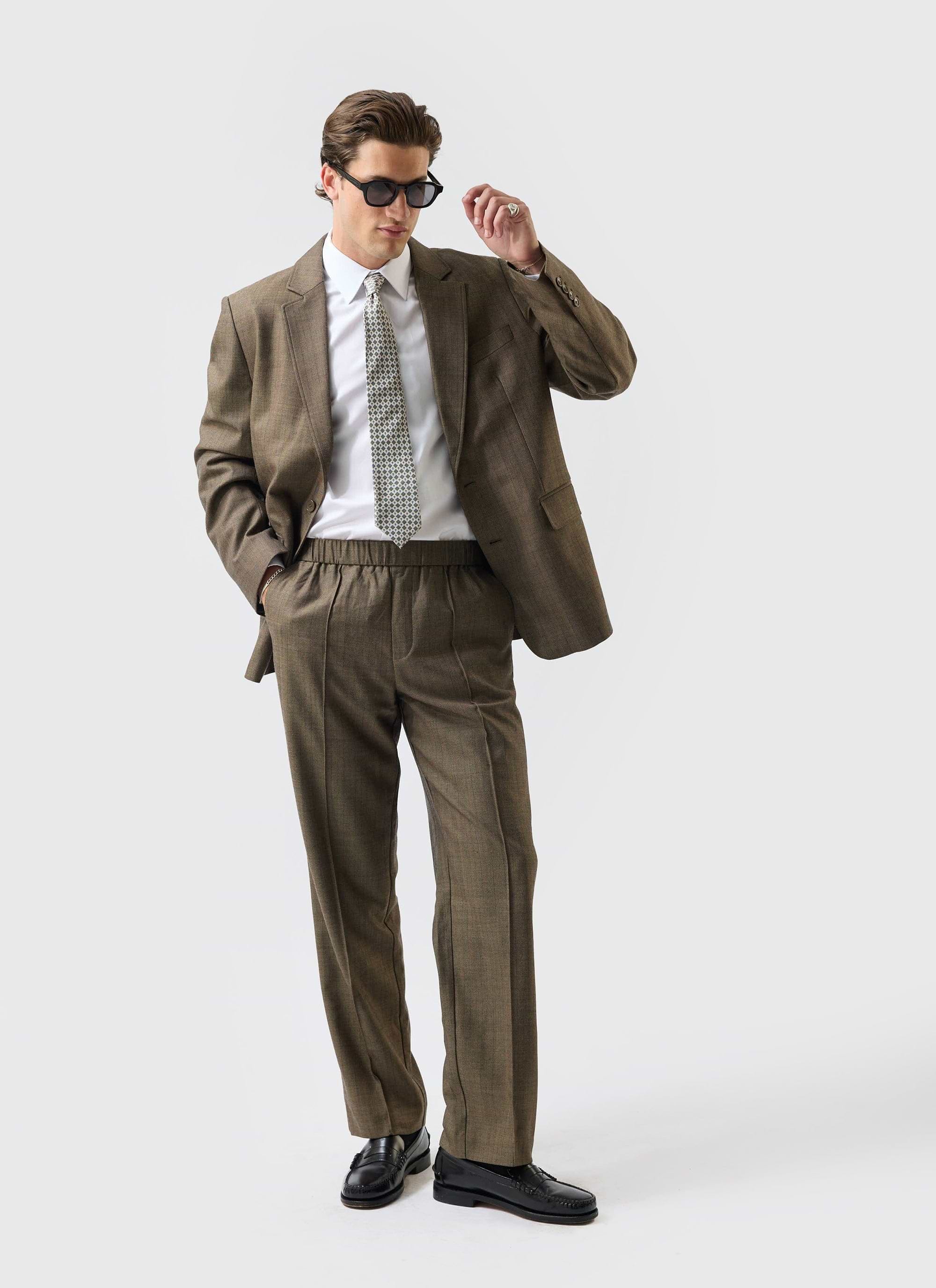 A model wears the Percival Grounds Tailored Blazer and matching trousers in oak herringbone wool, adjusting his sunglasses.