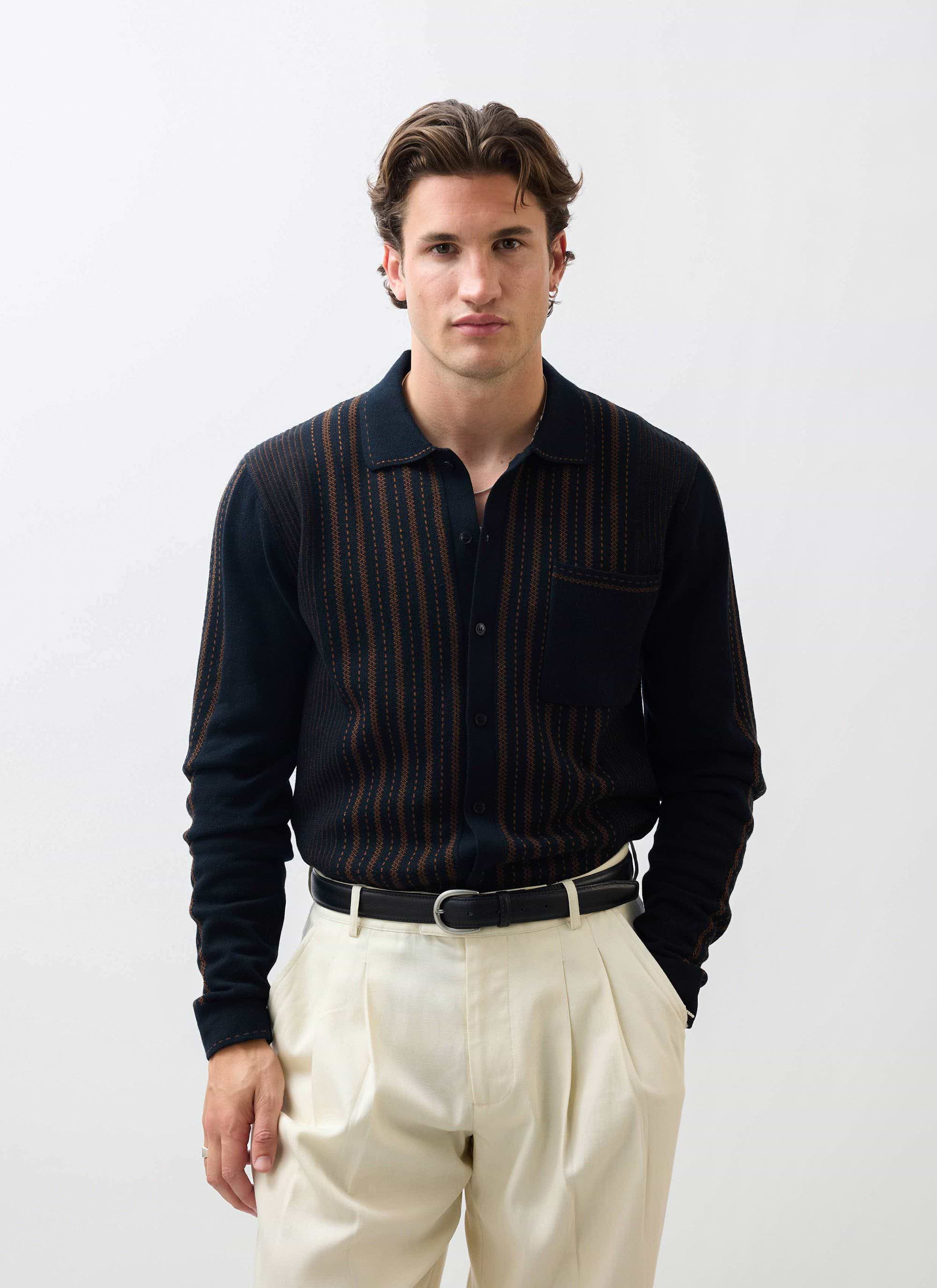 A model wears the Percival Appendix Long Sleeve Knitted Shirt in black with brown stripes, paired with cream trousers.