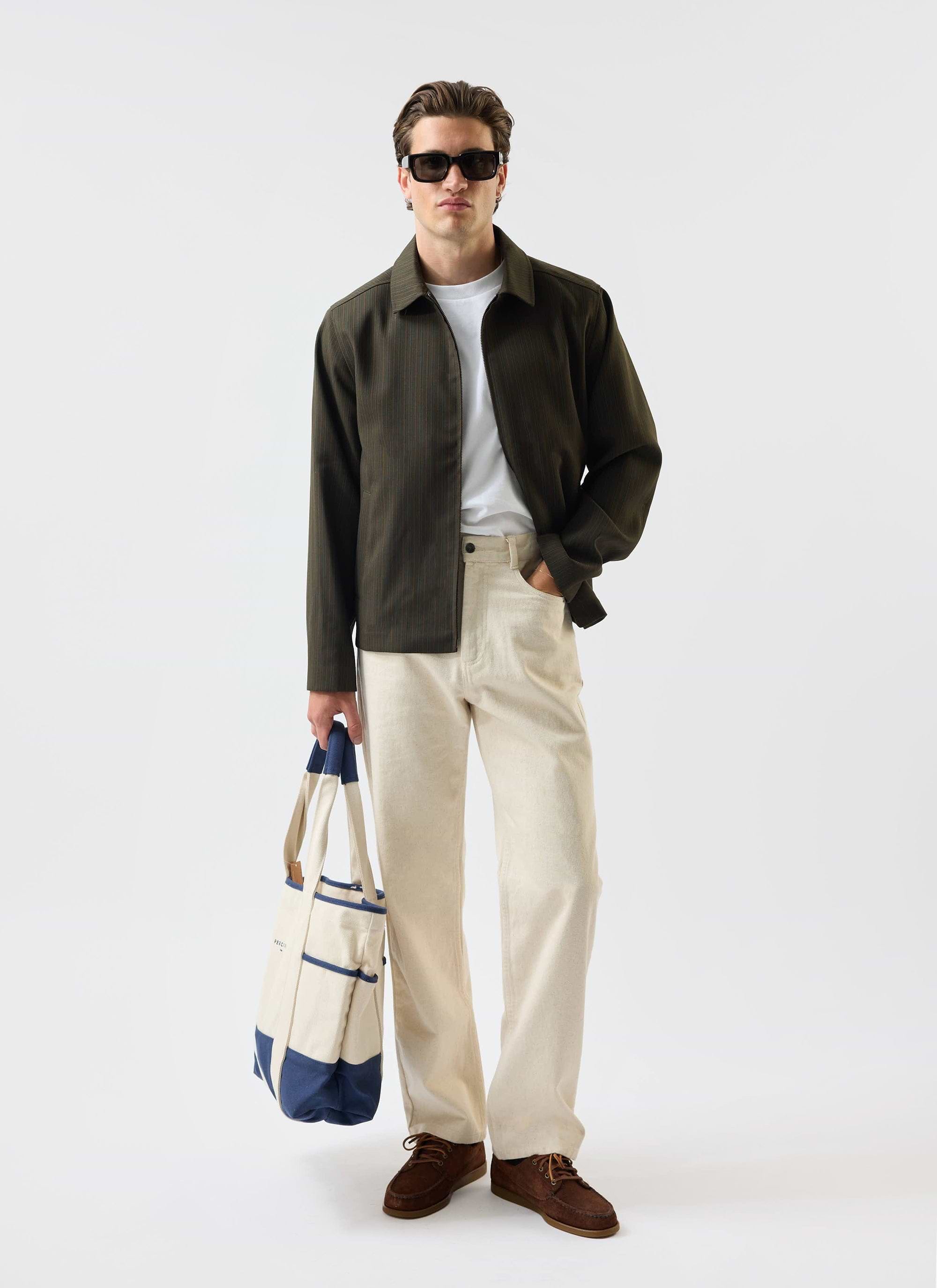 A model wears the Percival Chapter Zip Up Jacket in forest green, paired with cream trousers, sunglasses, and a tote bag.