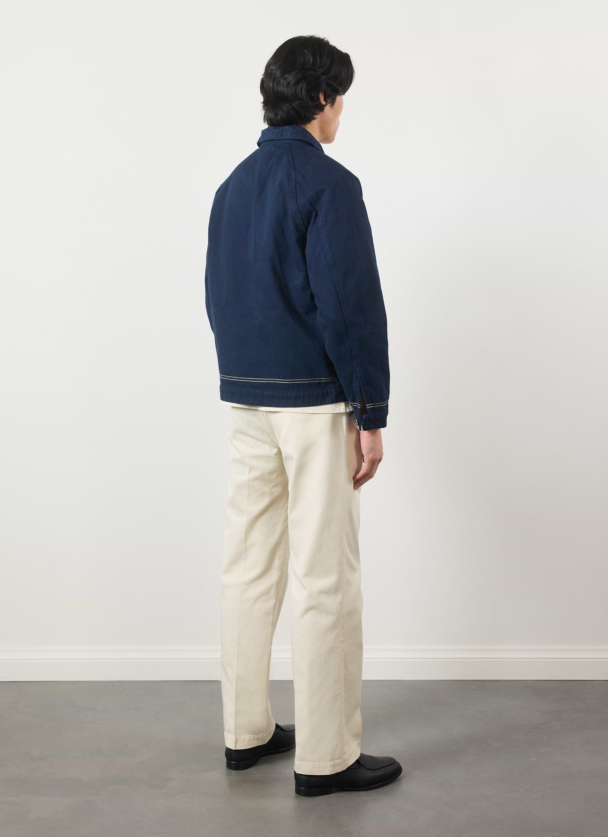 A model wears the Percival Calabria Harrington Jacket in navy with cream trousers, shot from the back against a white wall.