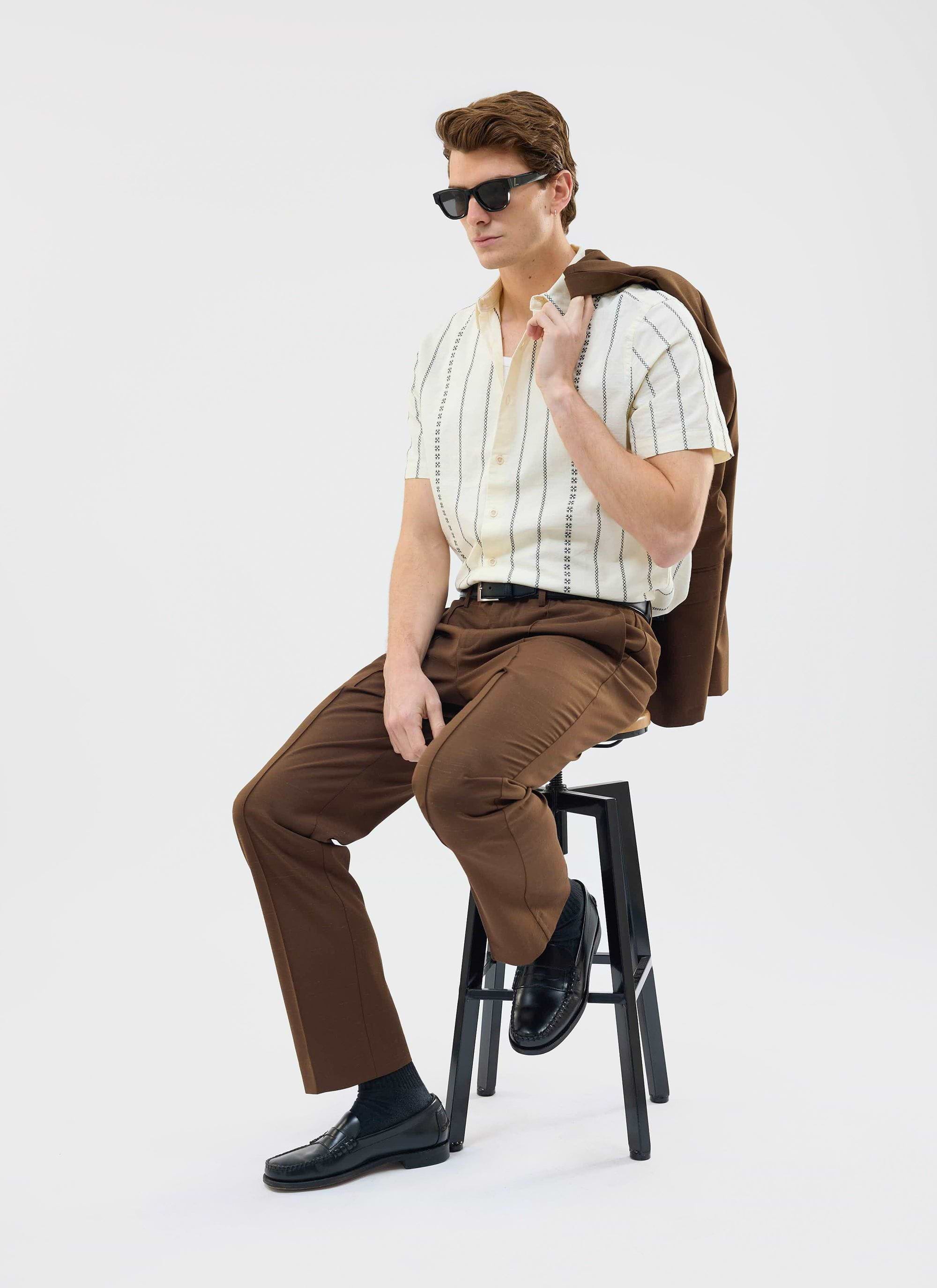A model poses on a stool wearing the Percival Cross Stitch Boxy Clerk Shirt in ecru with brown trousers and sunglasses.