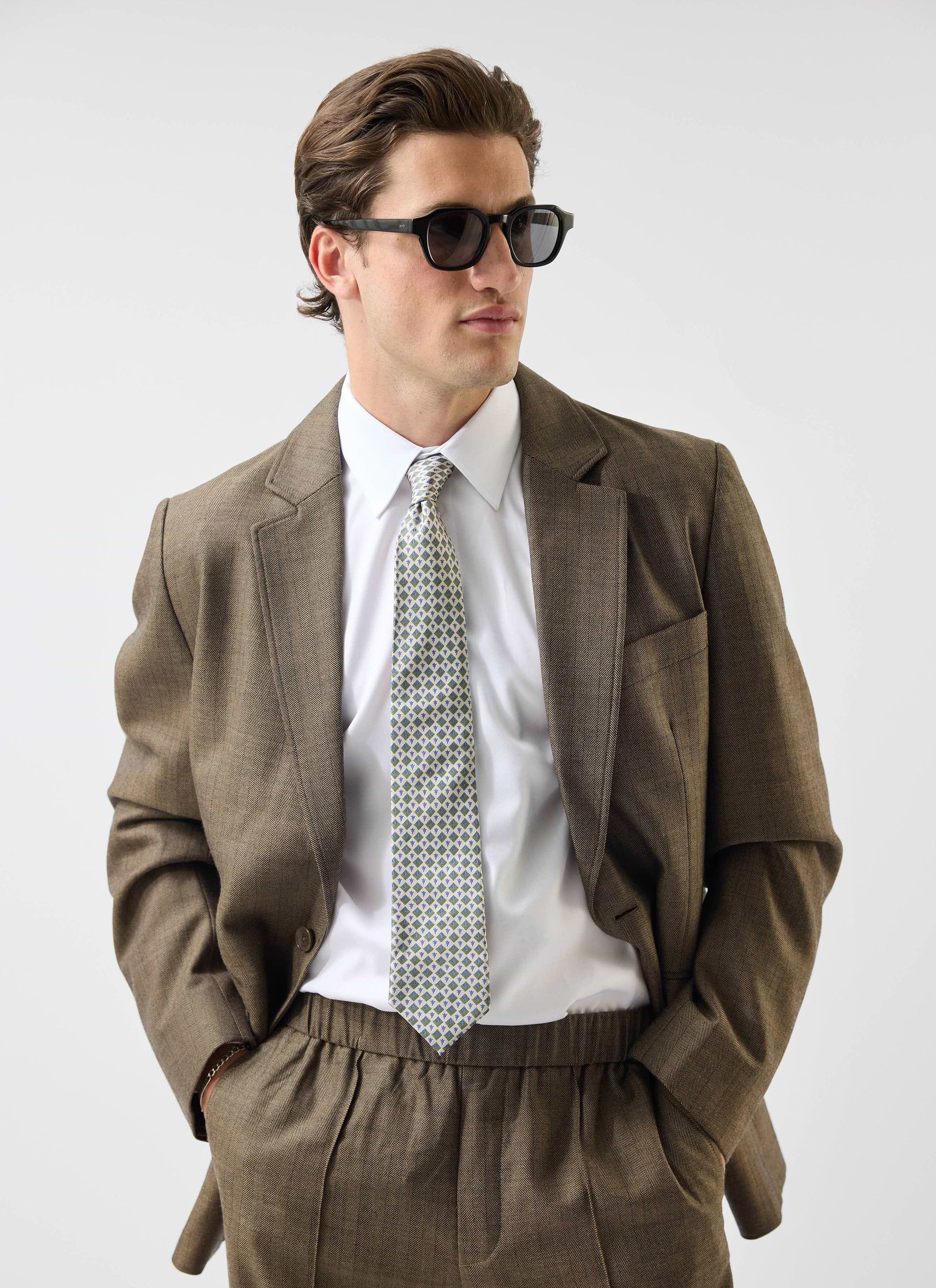 A man models the Percival Grounds Tailored Blazer in oak herringbone wool with a white shirt, tie and sunglasses.