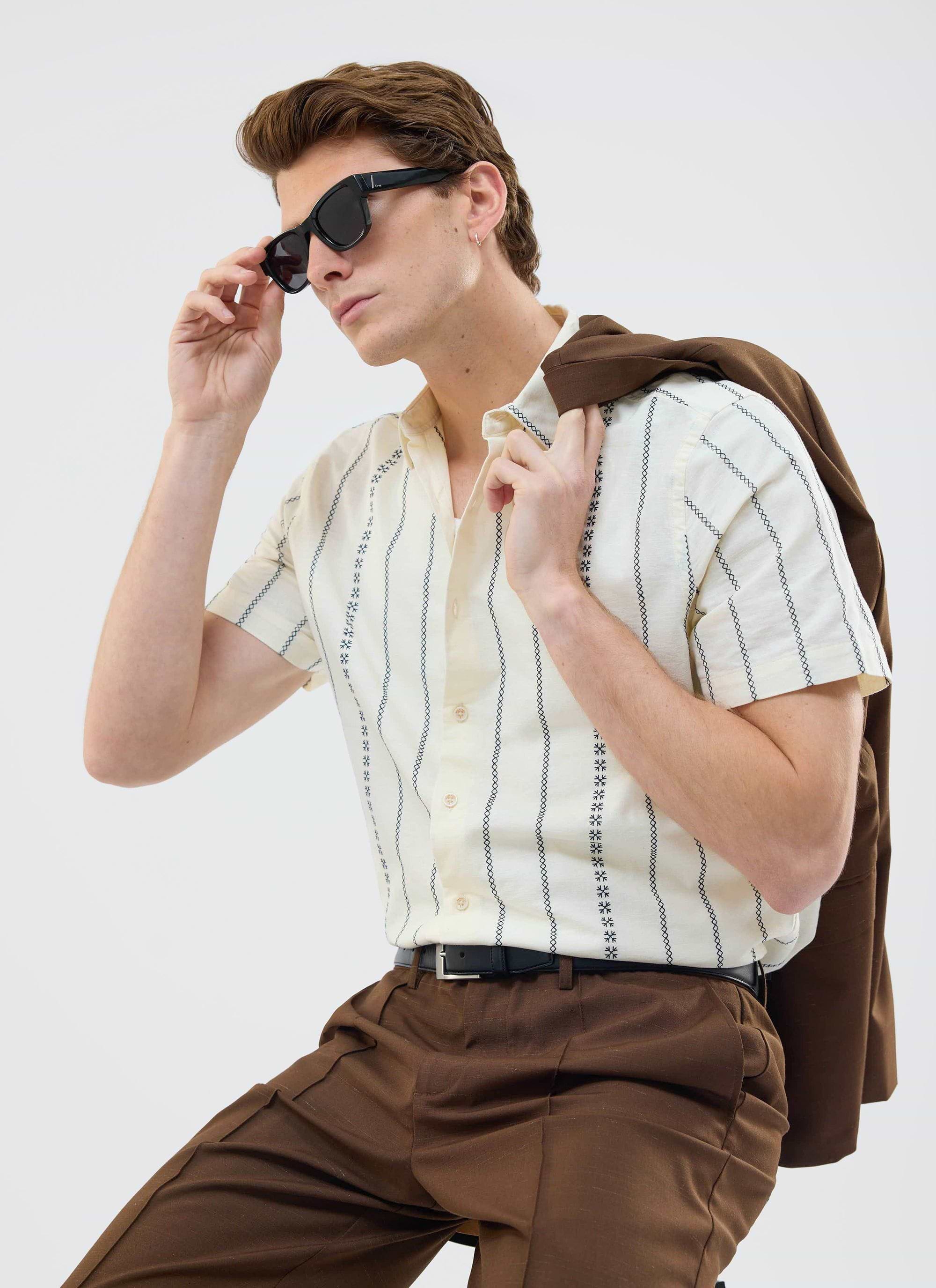 A model styled in the Percival Cross Stitch Boxy Clerk Shirt in ecru with brown trousers, adjusting his sunglasses.
