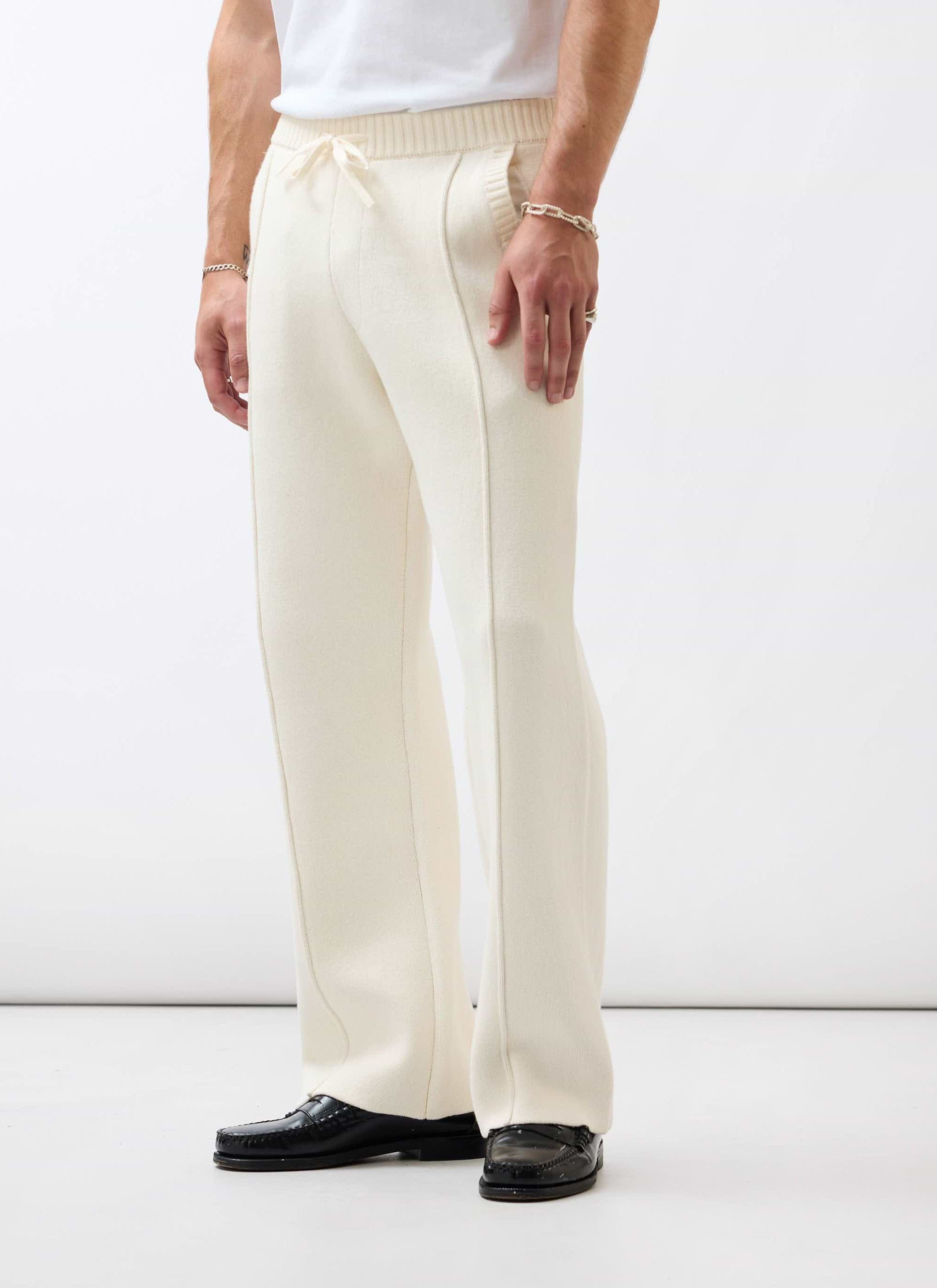 A man models the Percival Chronicle knitted trackpants in ecru, styled with black loafers against a white background.
