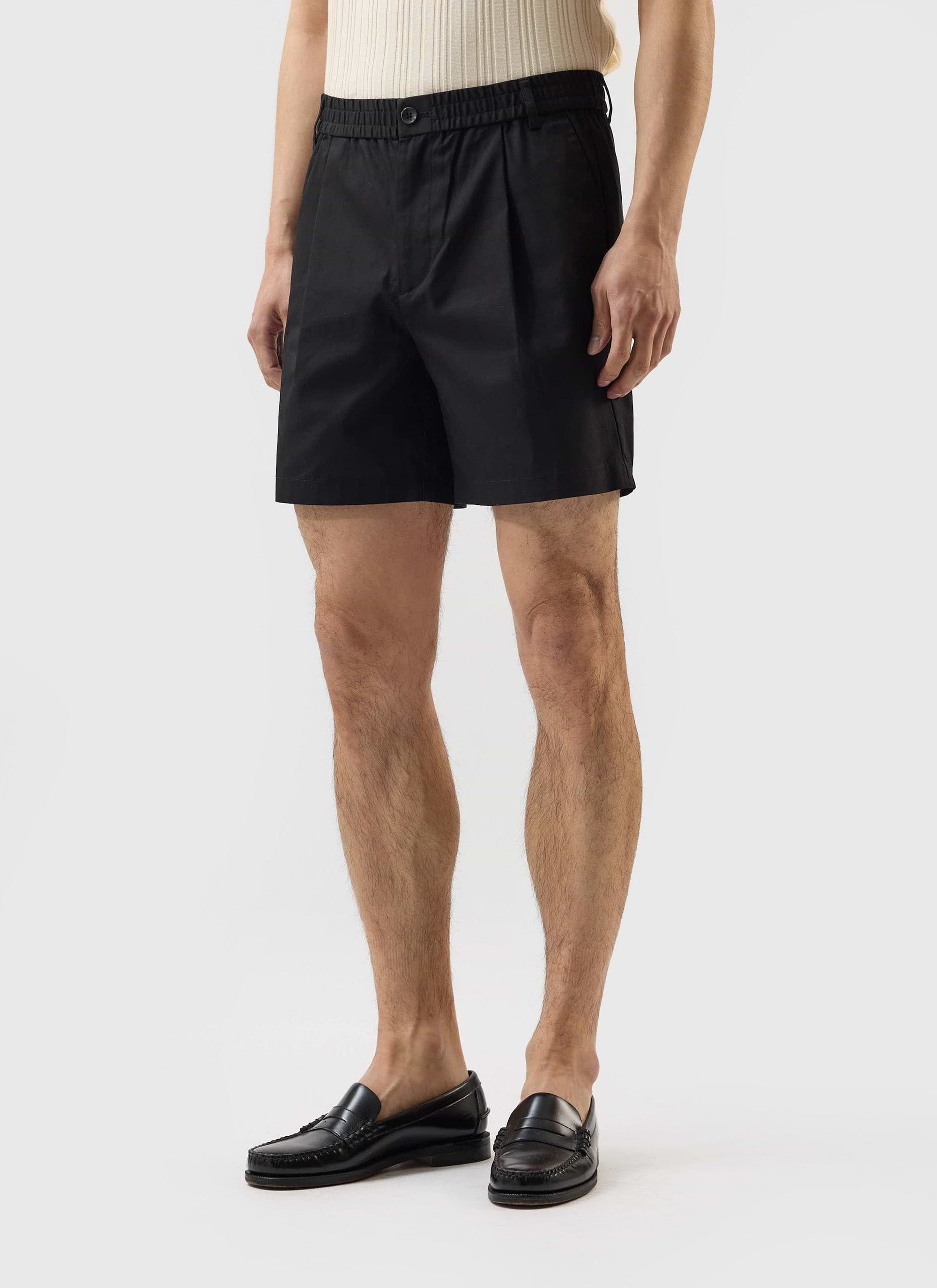 A model wears the black cotton Enzo Tailored Shorts by Percival, styled with a cream ribbed top and black loafers.
