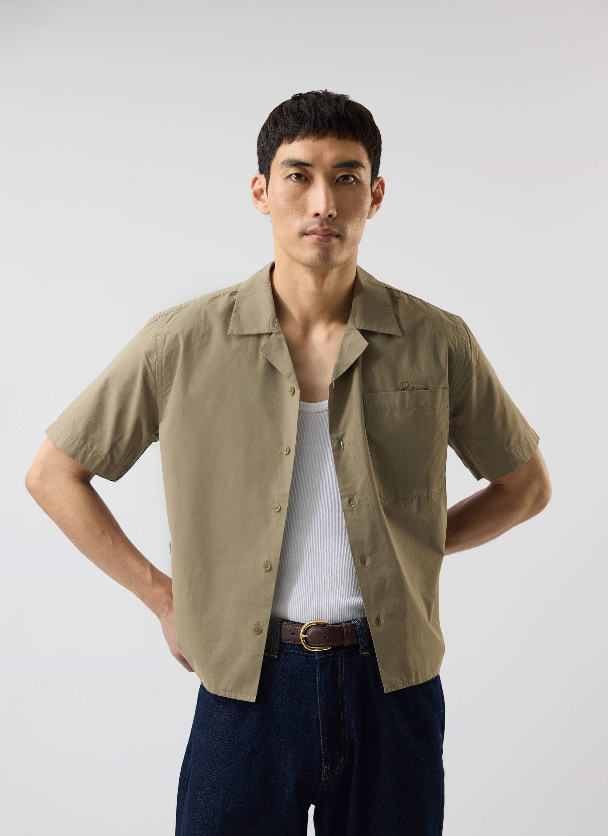 A model wears the Percival Boxy Cropped Cuban Collar Shirt in olive, styled open over a white vest with dark jeans.