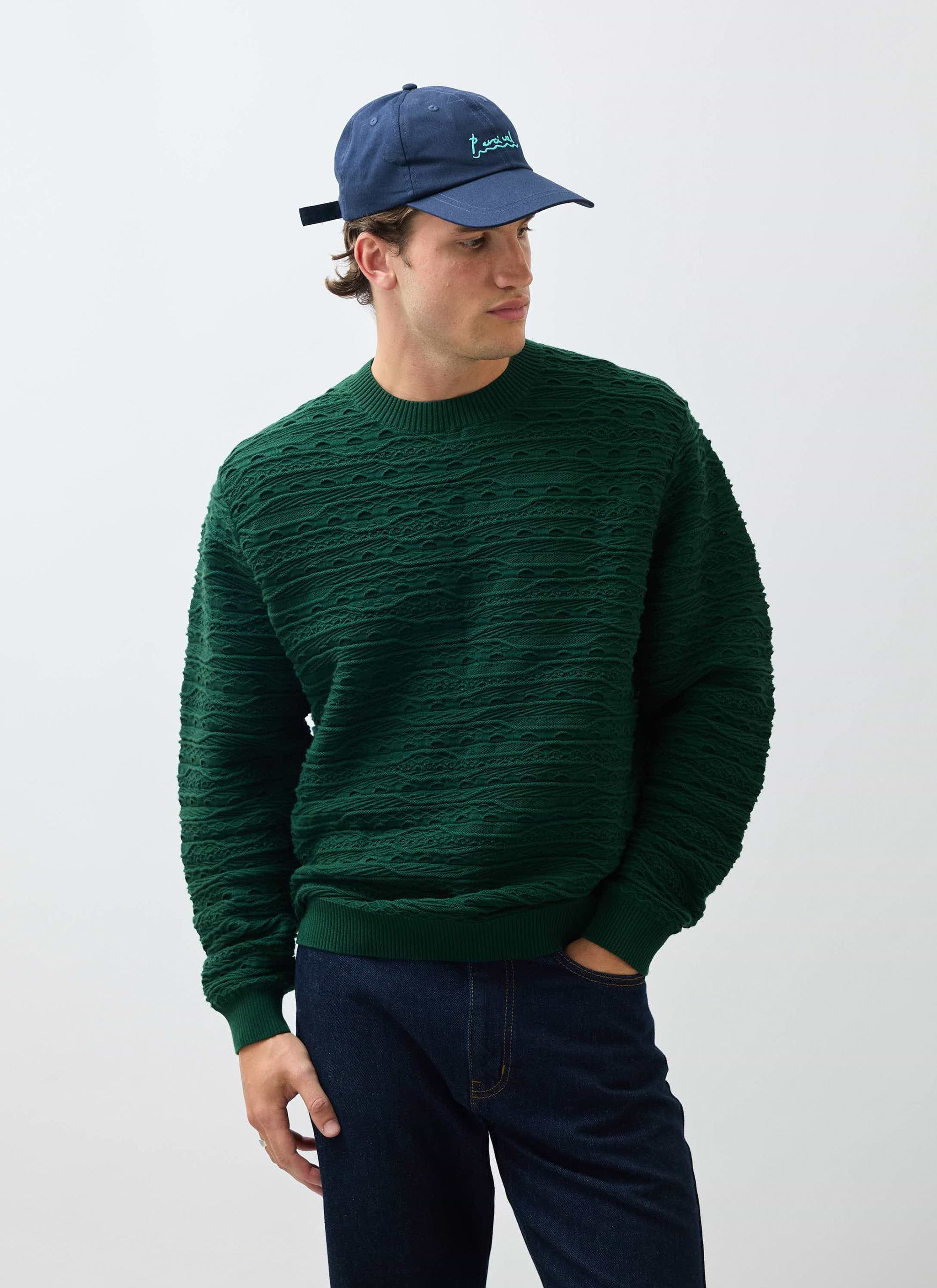 A model wears the textural Percival Tales Knitted Crew Neck Jumper in forest green, styled with dark jeans and a blue cap.