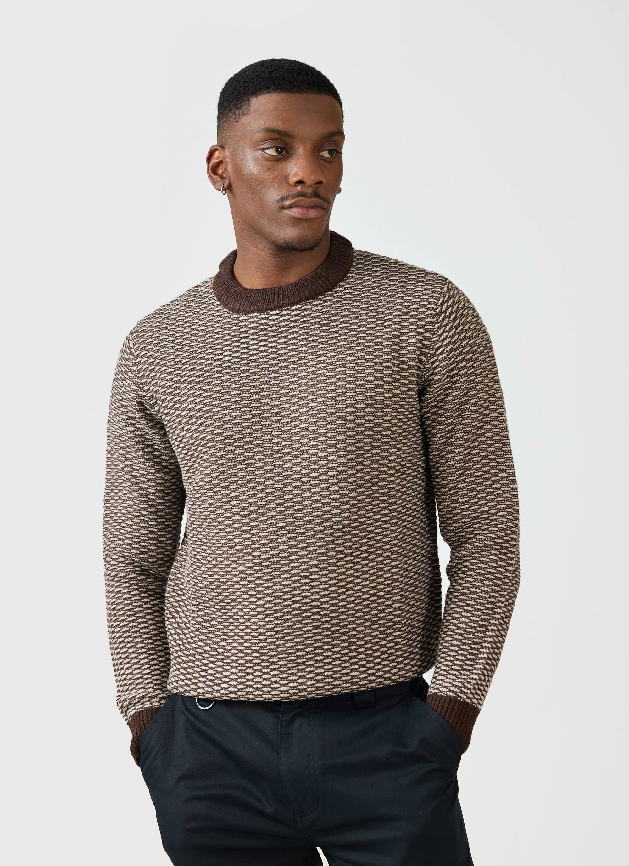 A man models the Percival Charlestown Knitted Jumper in a chocolate and beige textured knit, paired with black trousers.