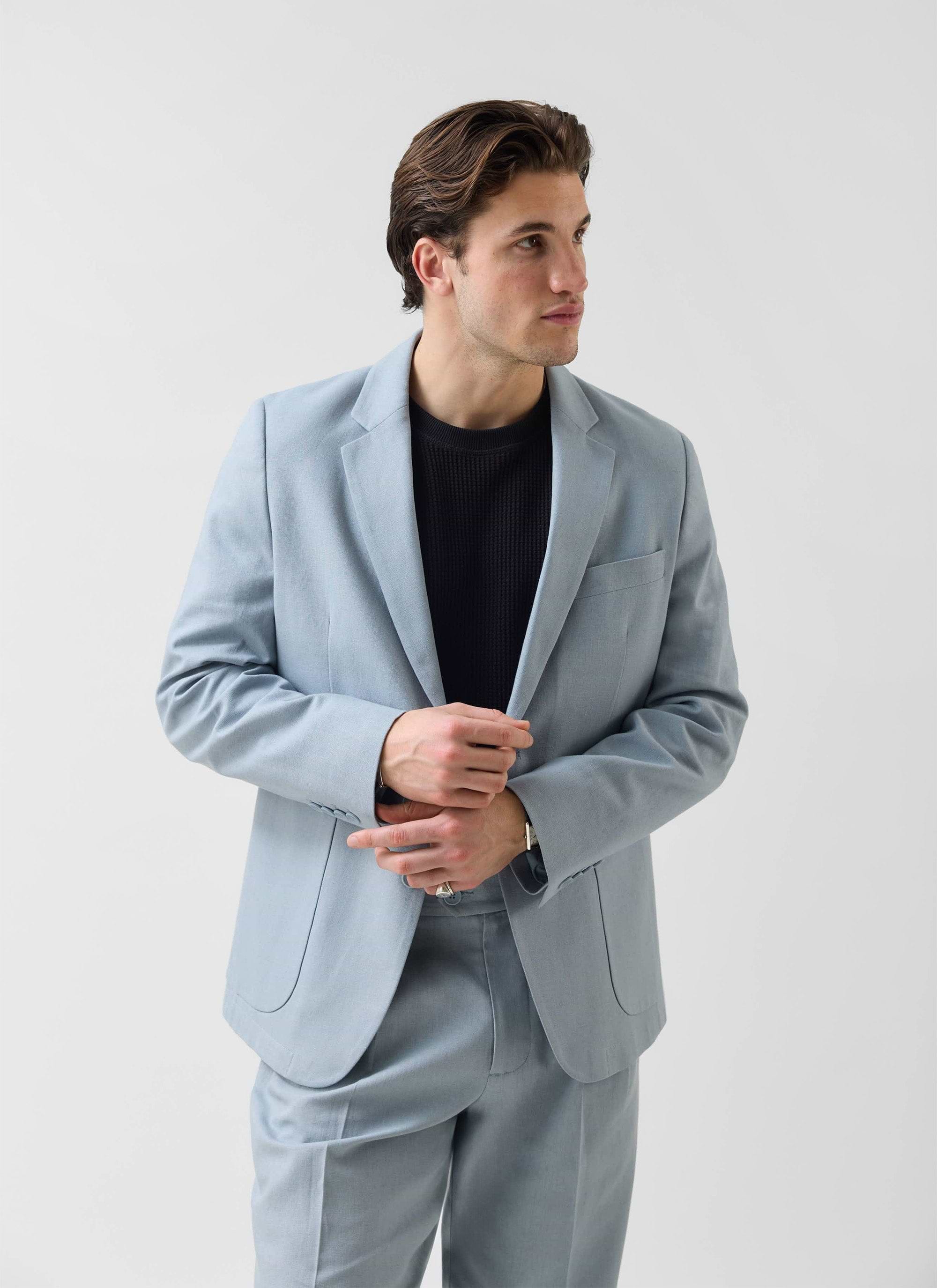 A man wears the Percival tailored linen blazer in light blue with matching trousers and a black knitted top.