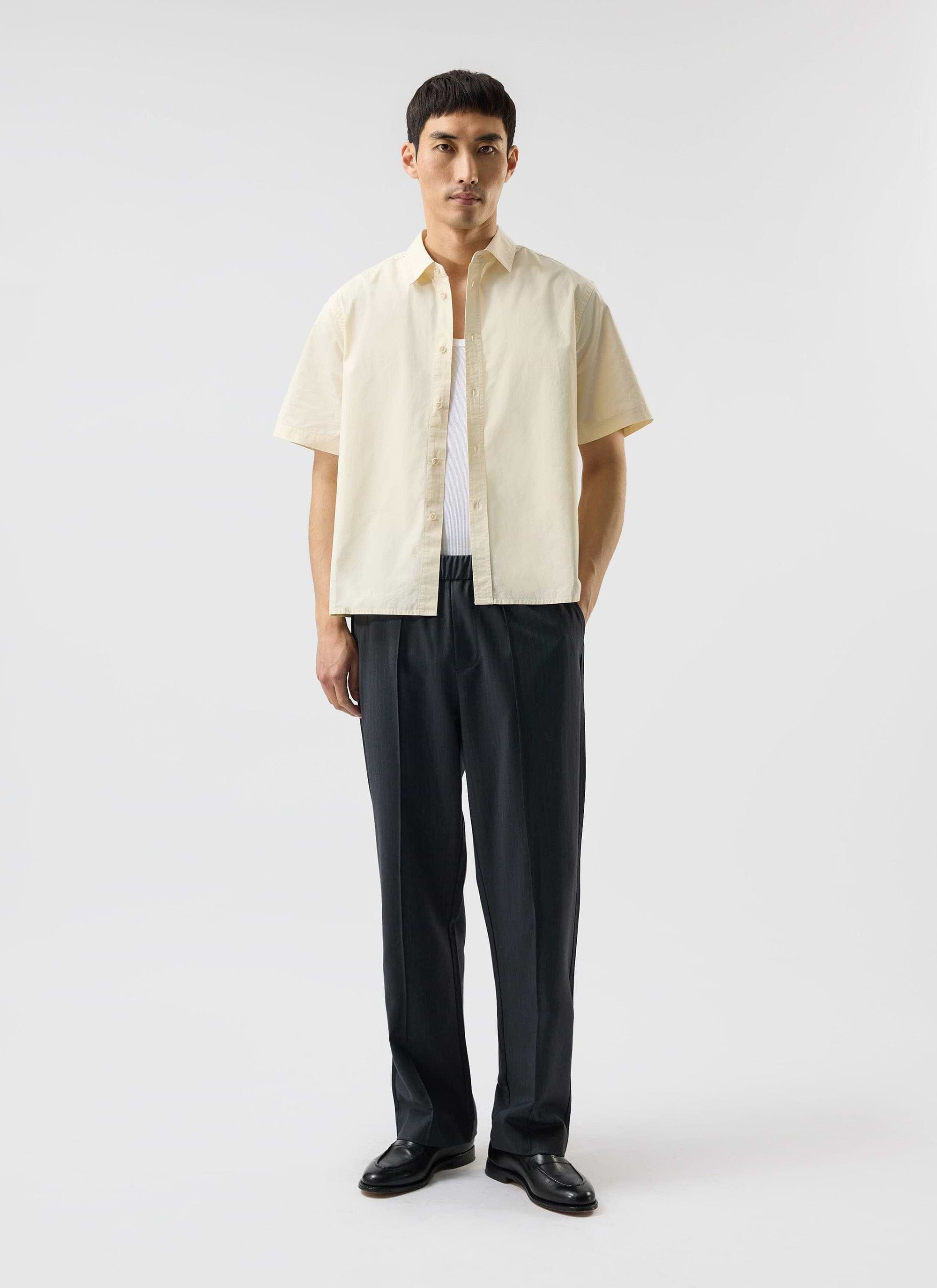 A model wears the Percival Boxy Cropped Clerk Shirt in ecru, styled open over a white vest and dark trousers.