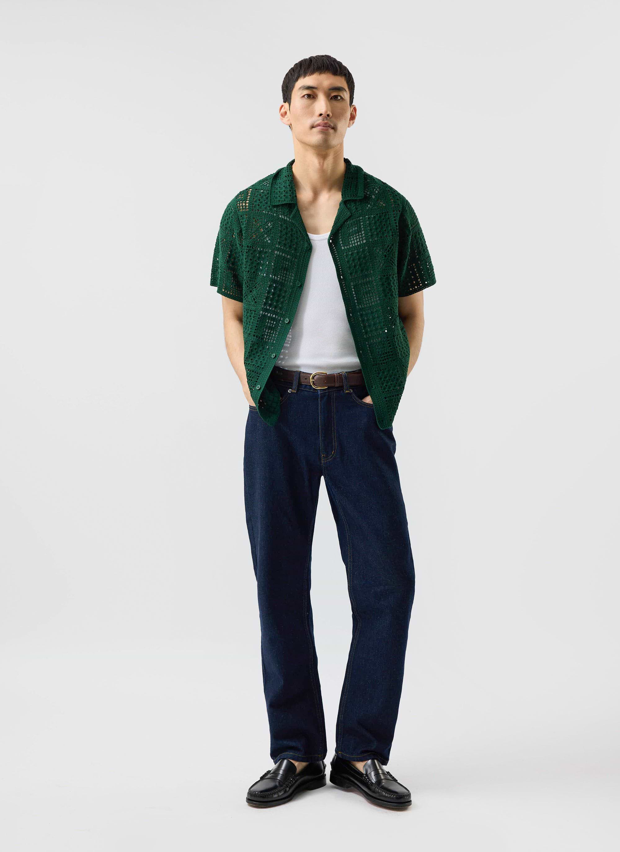 A man models the forest green Percival Crochet Groundsman Shirt, worn open over a white vest and styled with dark jeans.
