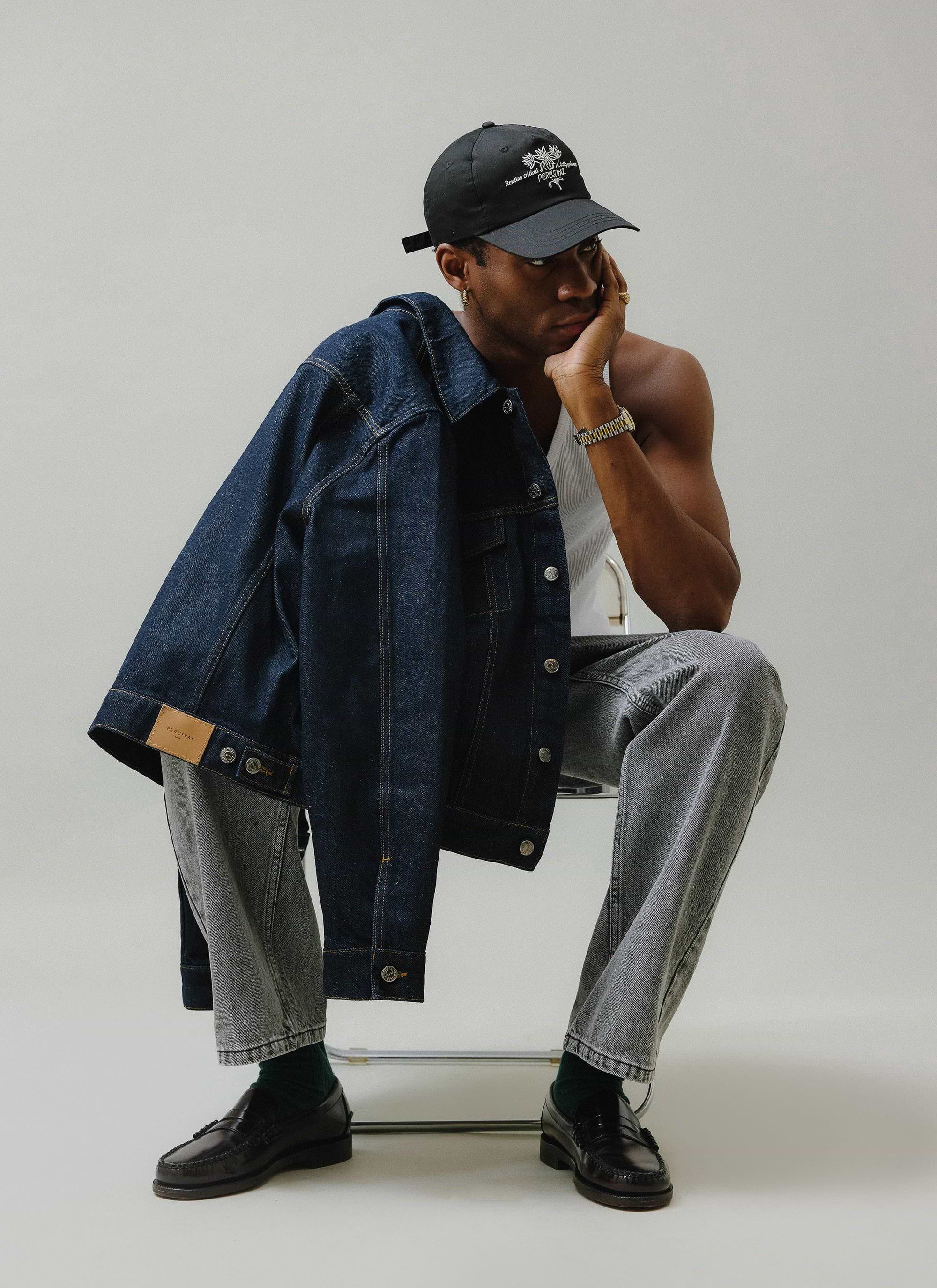 A model wearing Percival's warm grey wash wide leg jeans sits thoughtfully with a dark denim jacket over his shoulder.