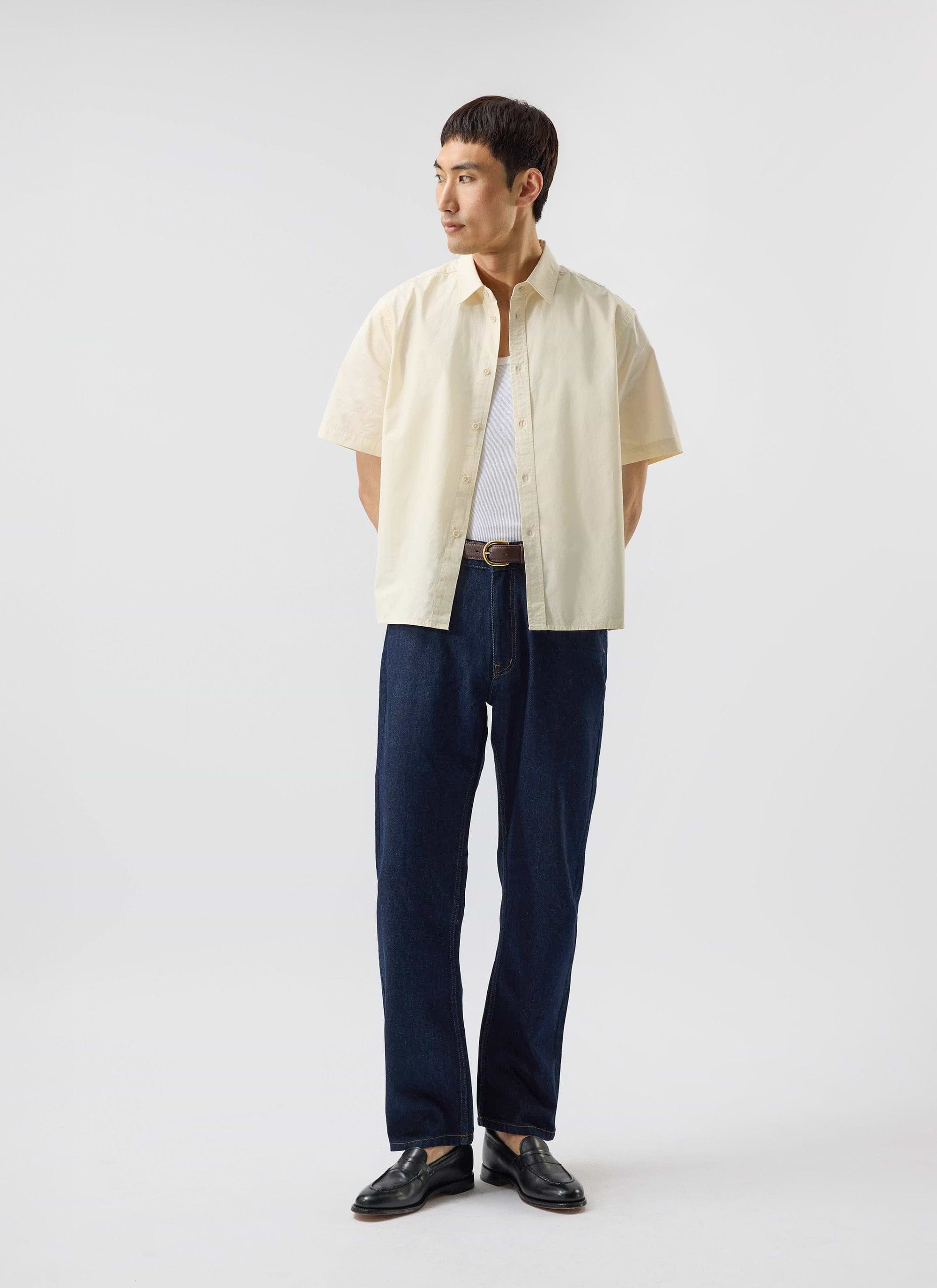 A man models the Percival Boxy Cropped Clerk Shirt in ecru, layered over a white vest and styled with dark denim jeans.