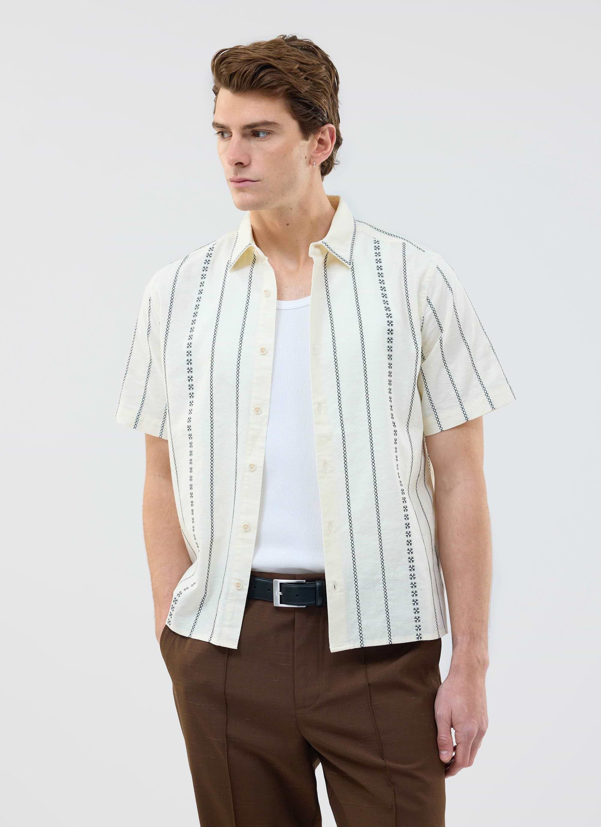 A model wears the Percival Cross Stitch Boxy Clerk Shirt in ecru, styled open over a white vest with brown trousers.