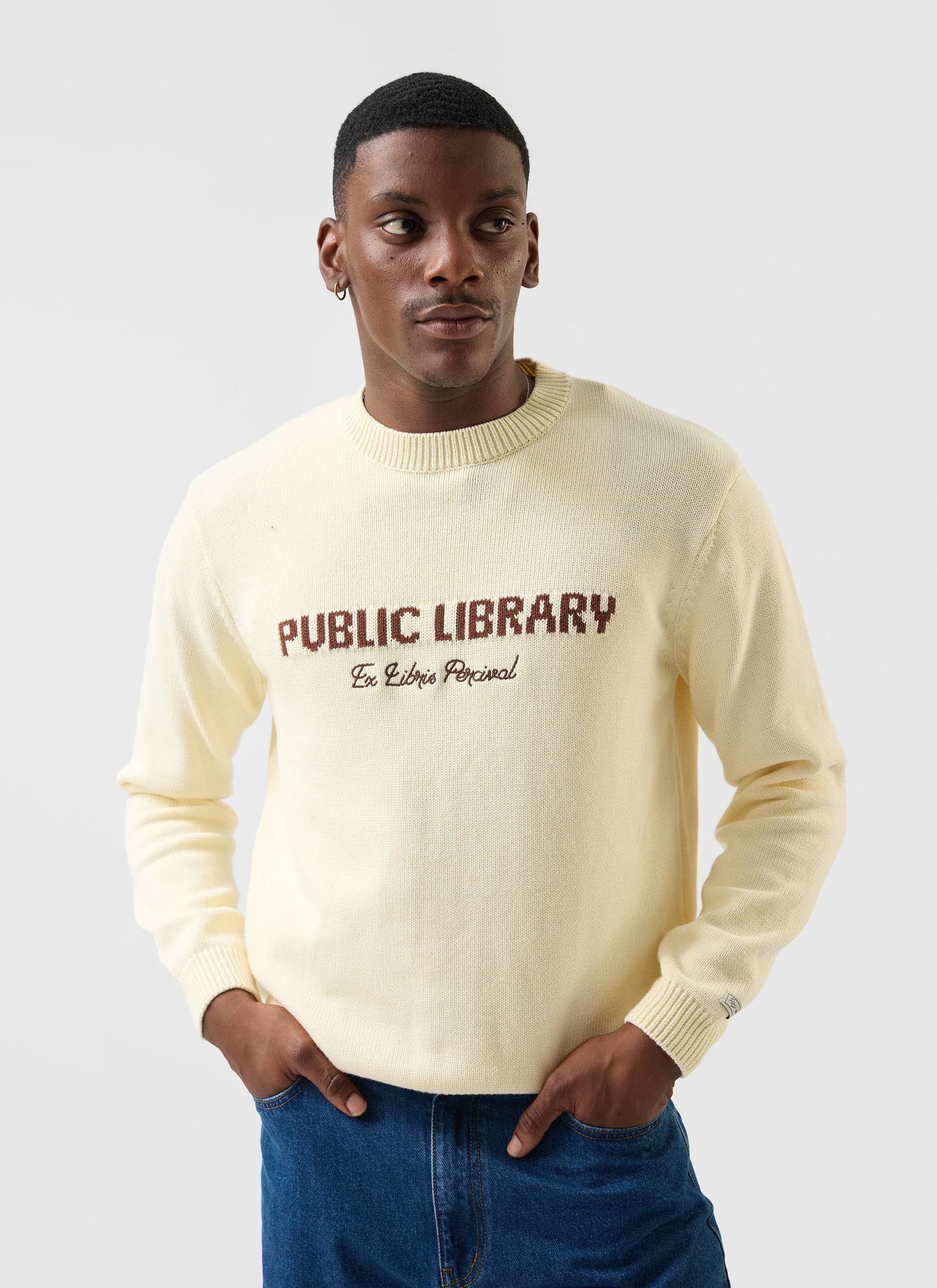 A model wears the Percival Public Library knitted jumper in ecru cream, featuring brown chest embroidery.
