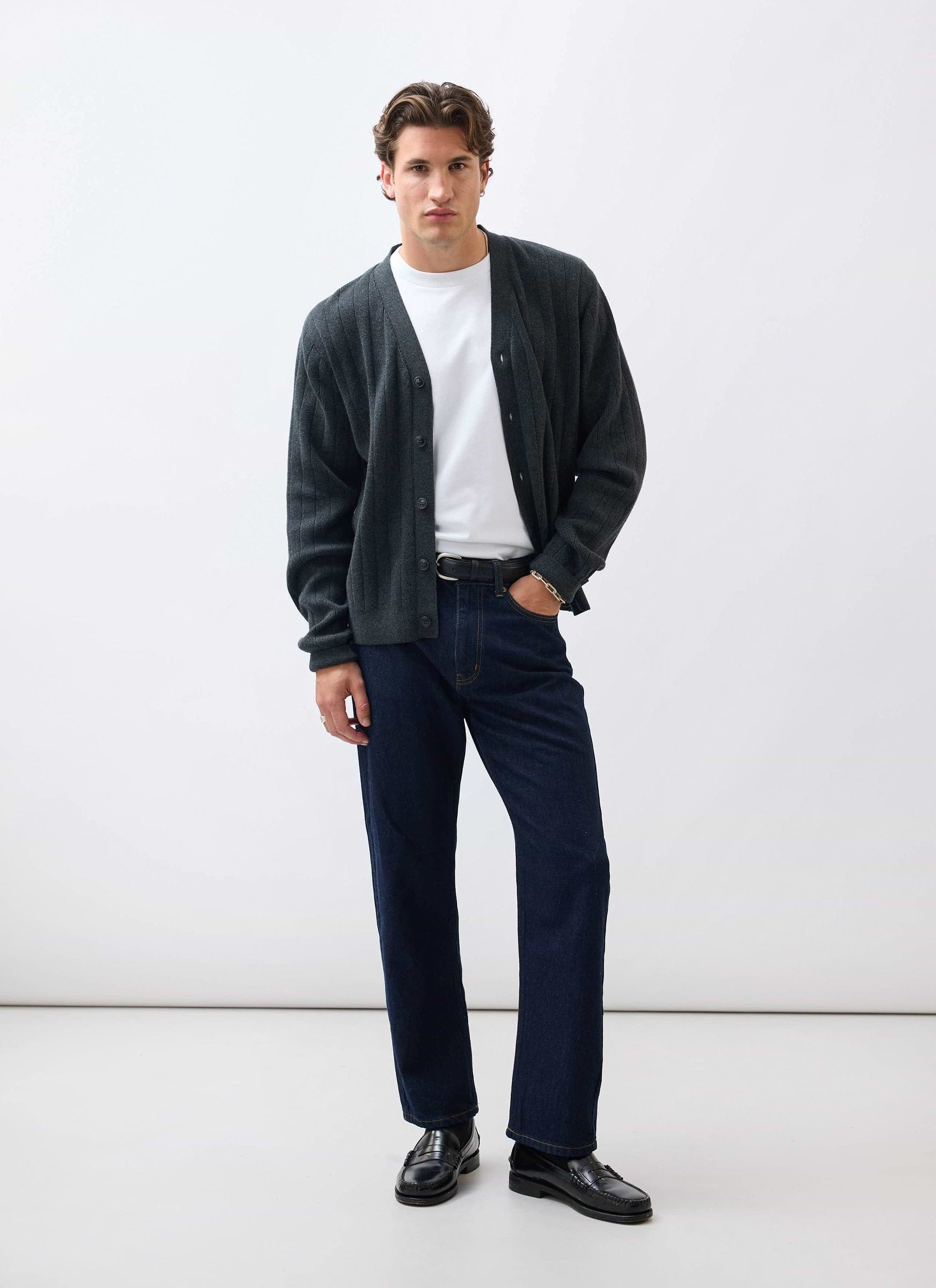 A man models the Percival Editor Cardigan in grey, styled with a white t-shirt, dark jeans, and black loafers.