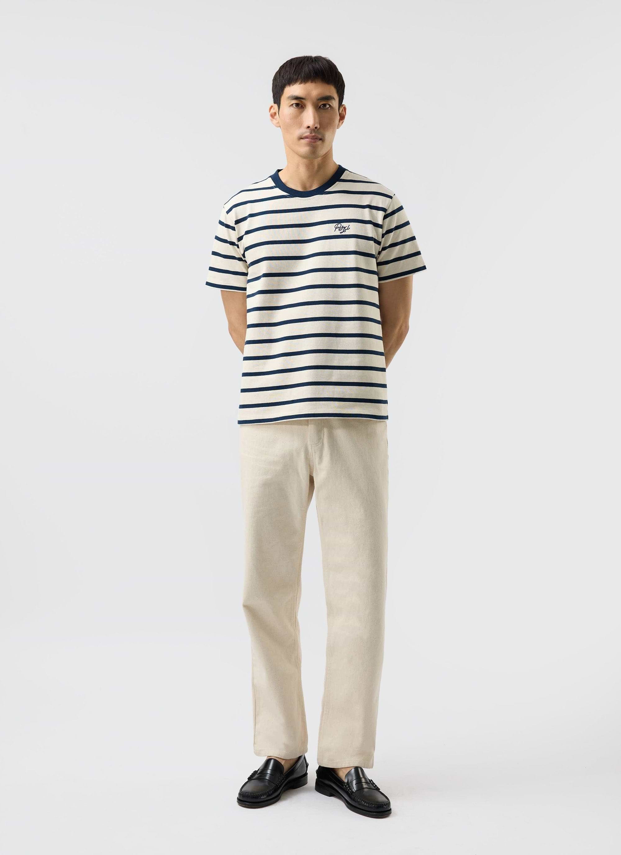 A man models the Percival Breton Stripe T-shirt in ecru with navy, paired with ecru trousers and black loafers.