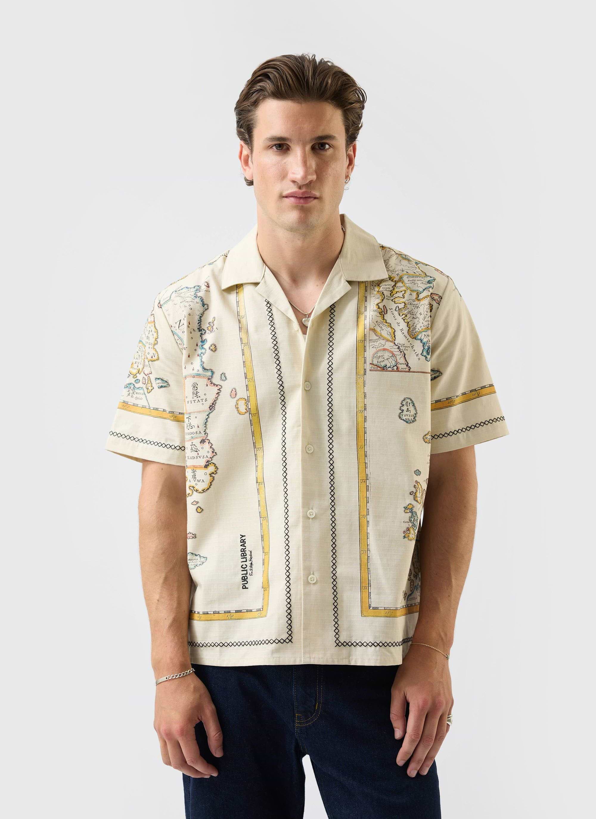 A man models the ecru Ex Libris Cuban Shirt from Percival, showing the detailed world map artwork and embroidery.