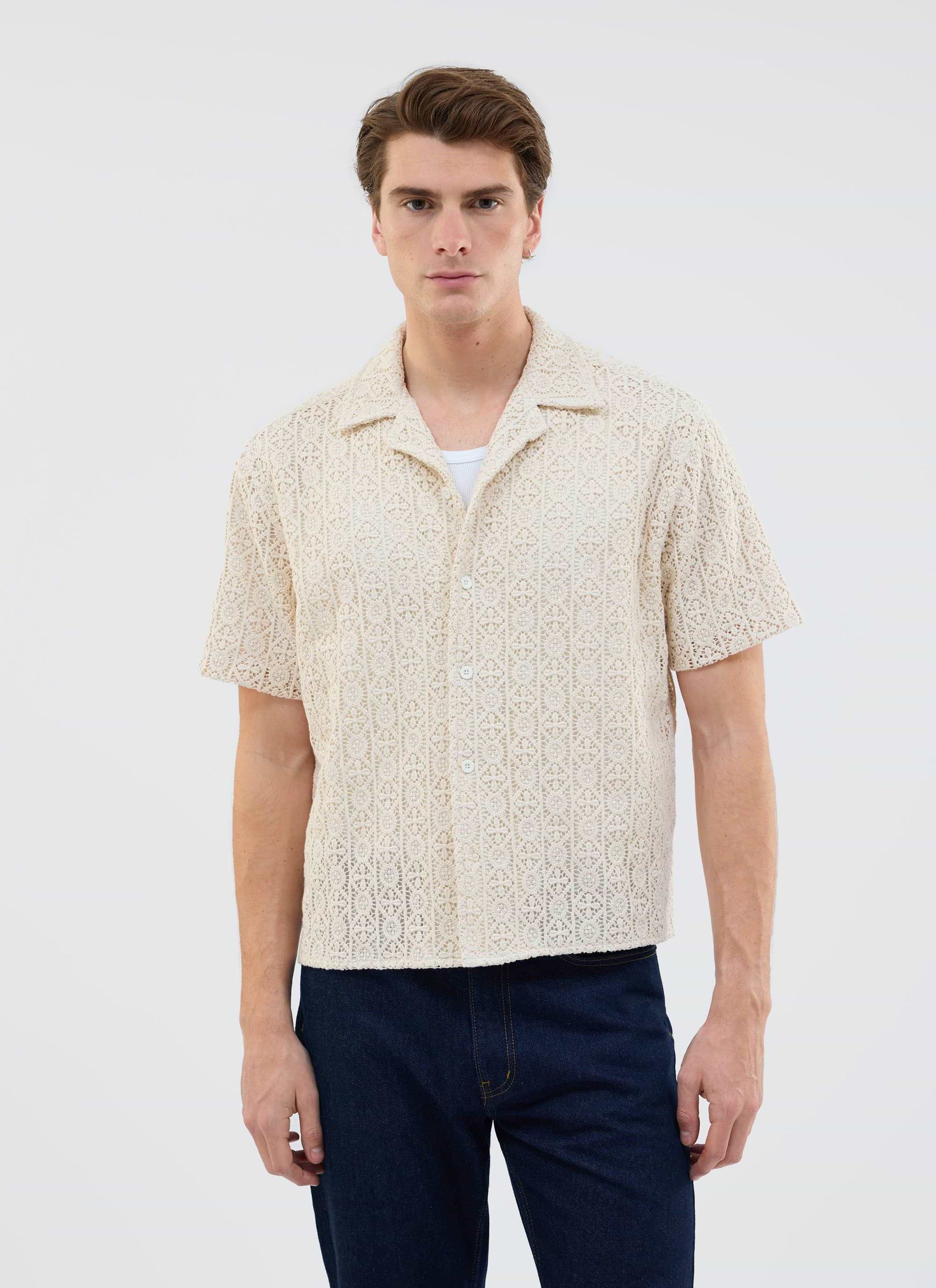 A man models the Percival Elio Crochet Cuban Shirt in ecru, styled over a white vest and paired with dark wash jeans.