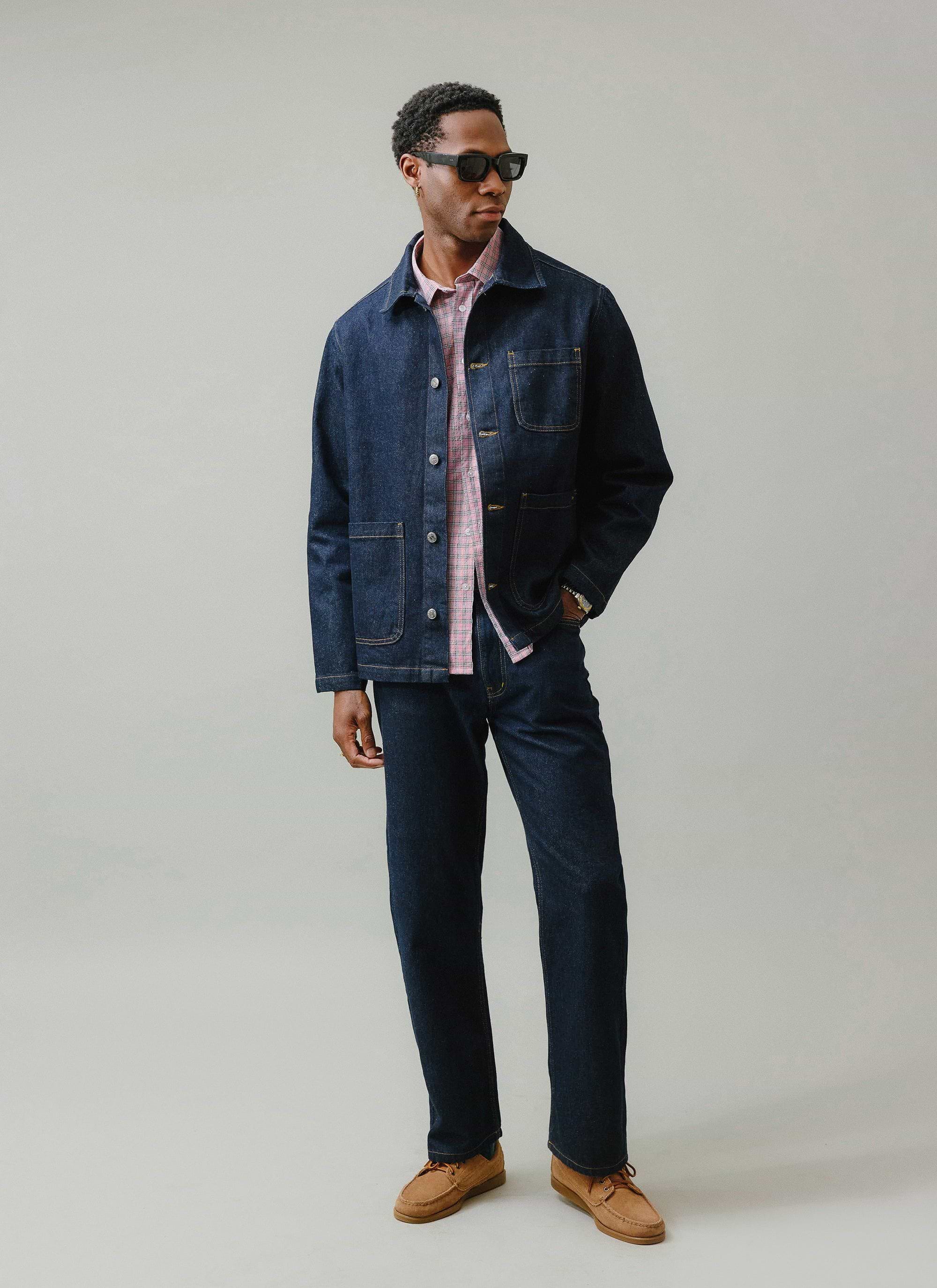 A man models the Percival Gove Chore Jacket in dark indigo with matching denim jeans and a pink checked shirt.