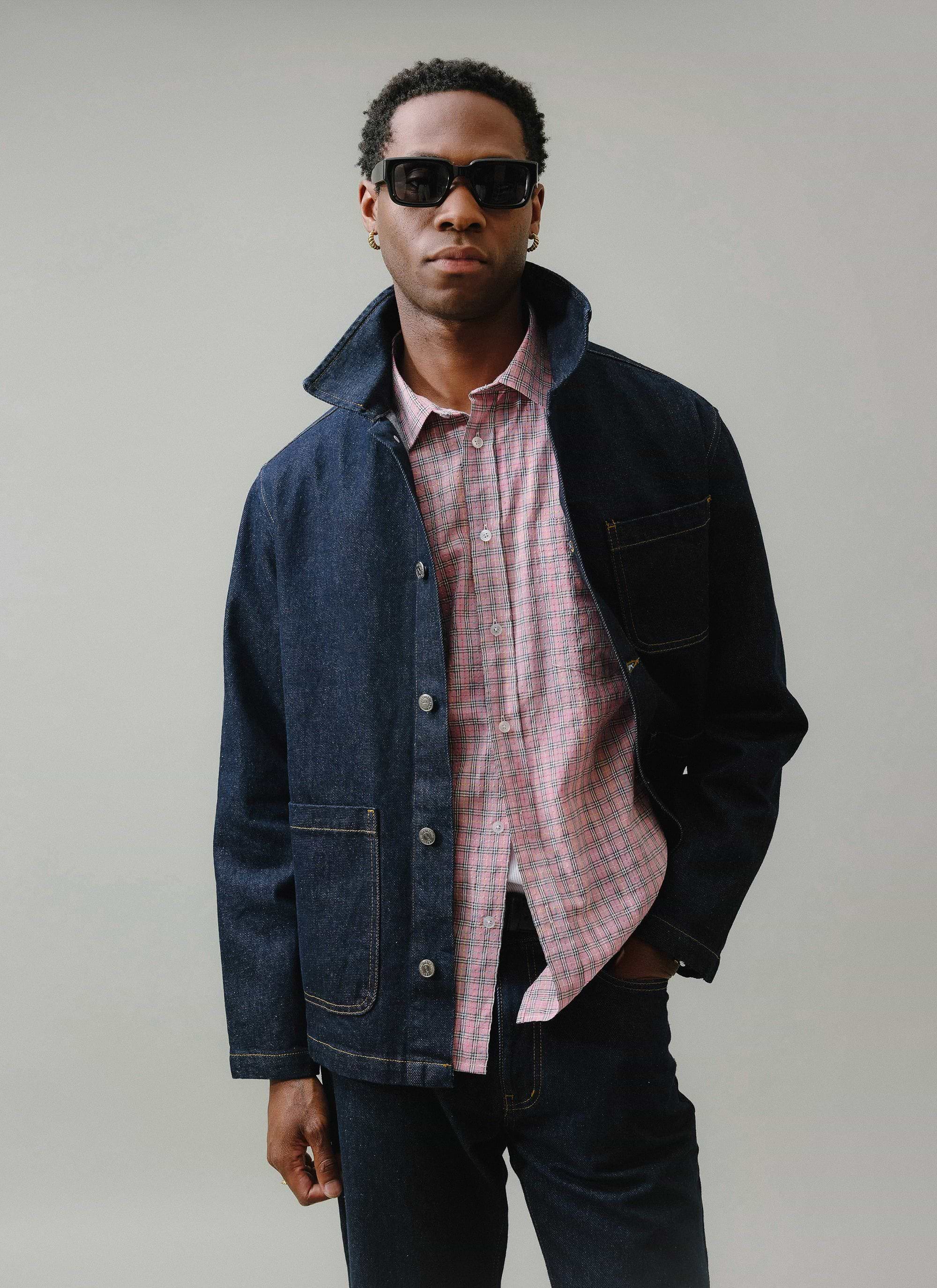 A model wears the Percival Gove Chore Jacket in dark indigo, styled open over a pink checked shirt with matching denim jeans.