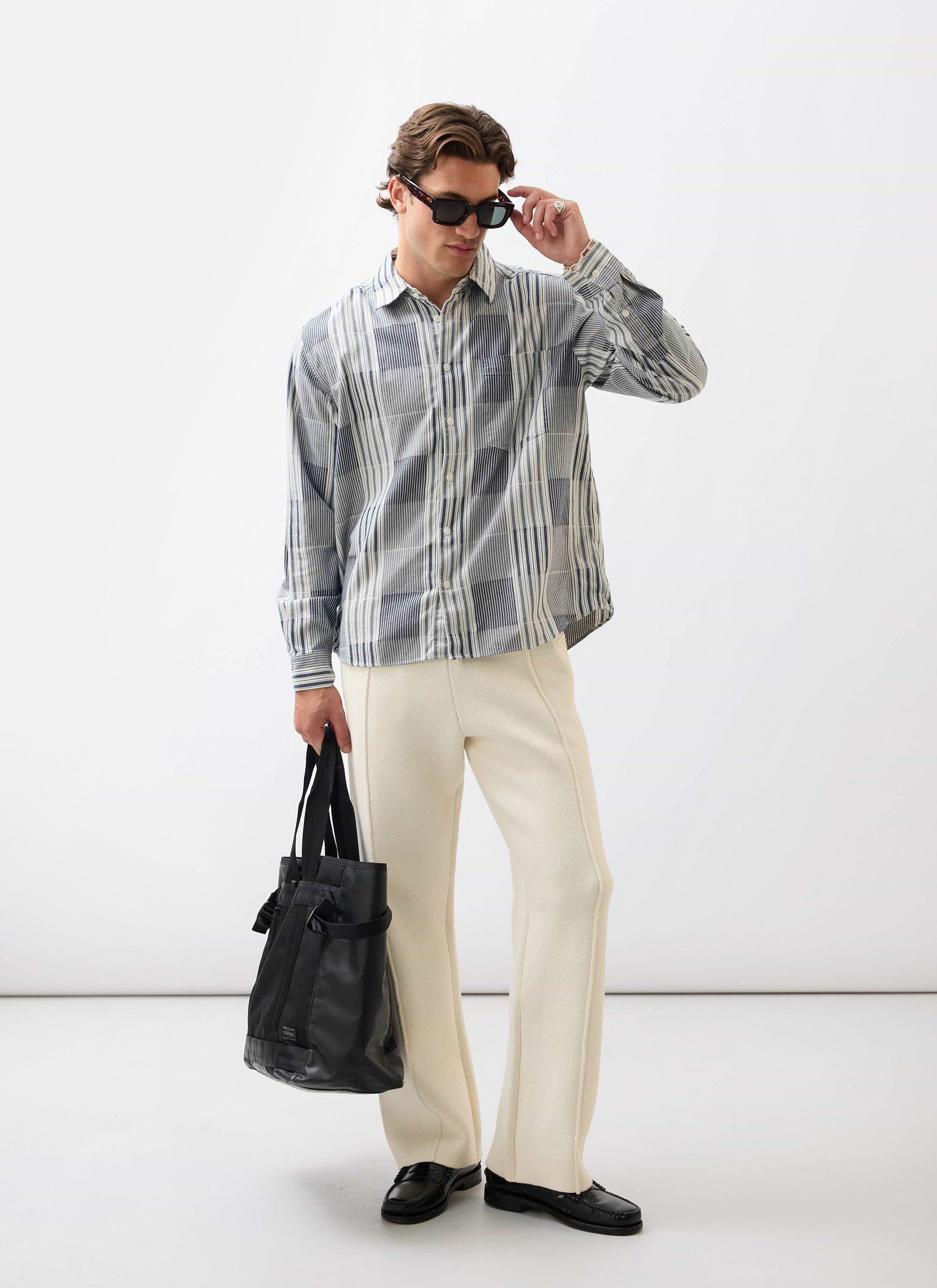A model wears the Percival Epigraph Patchwork Pearce shirt in blue with cream trousers, holding a black bag and sunglasses.
