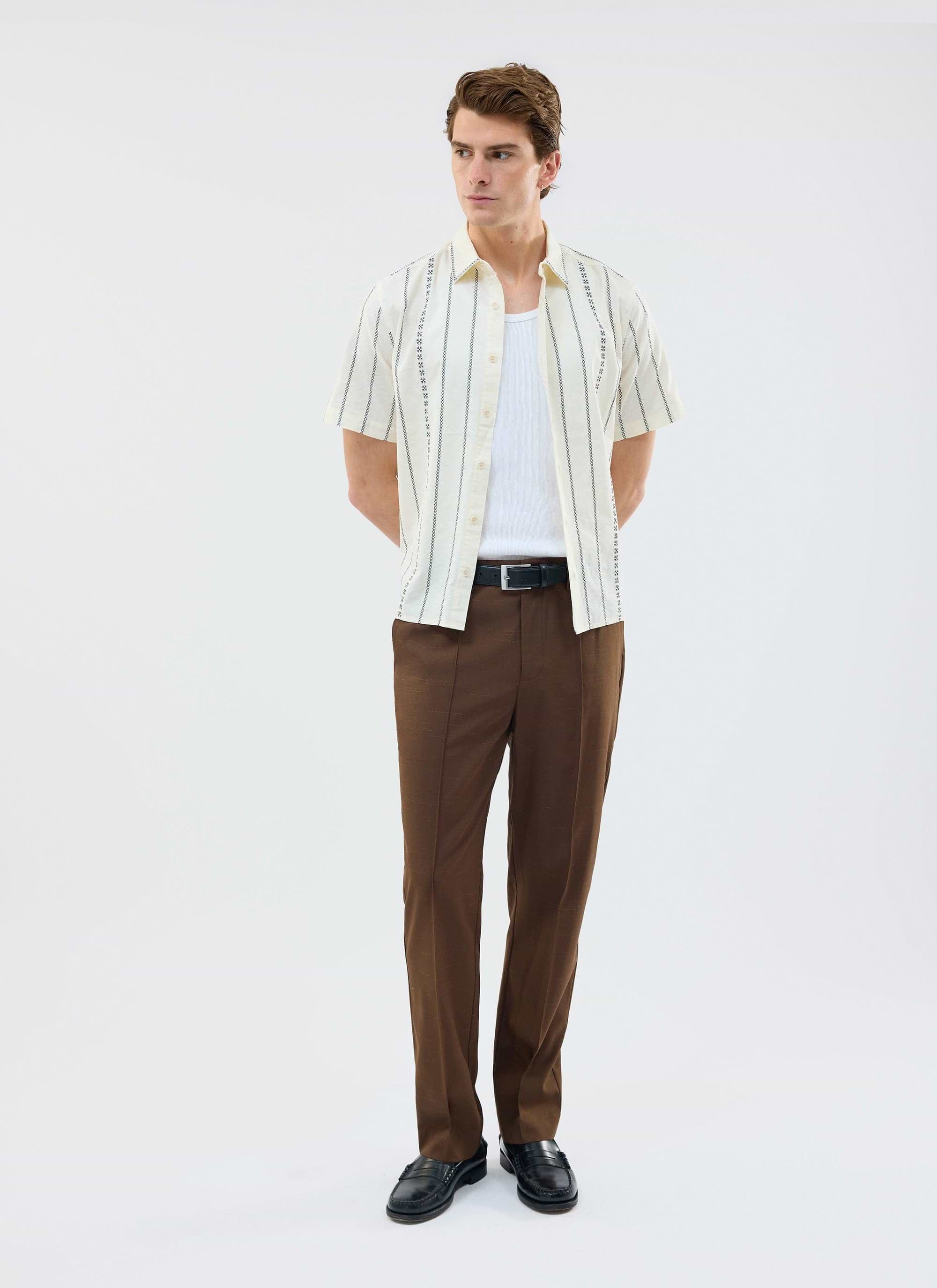 A man models the Percival Cross Stitch Boxy Clerk Shirt in ecru, worn open over a white vest with brown trousers.