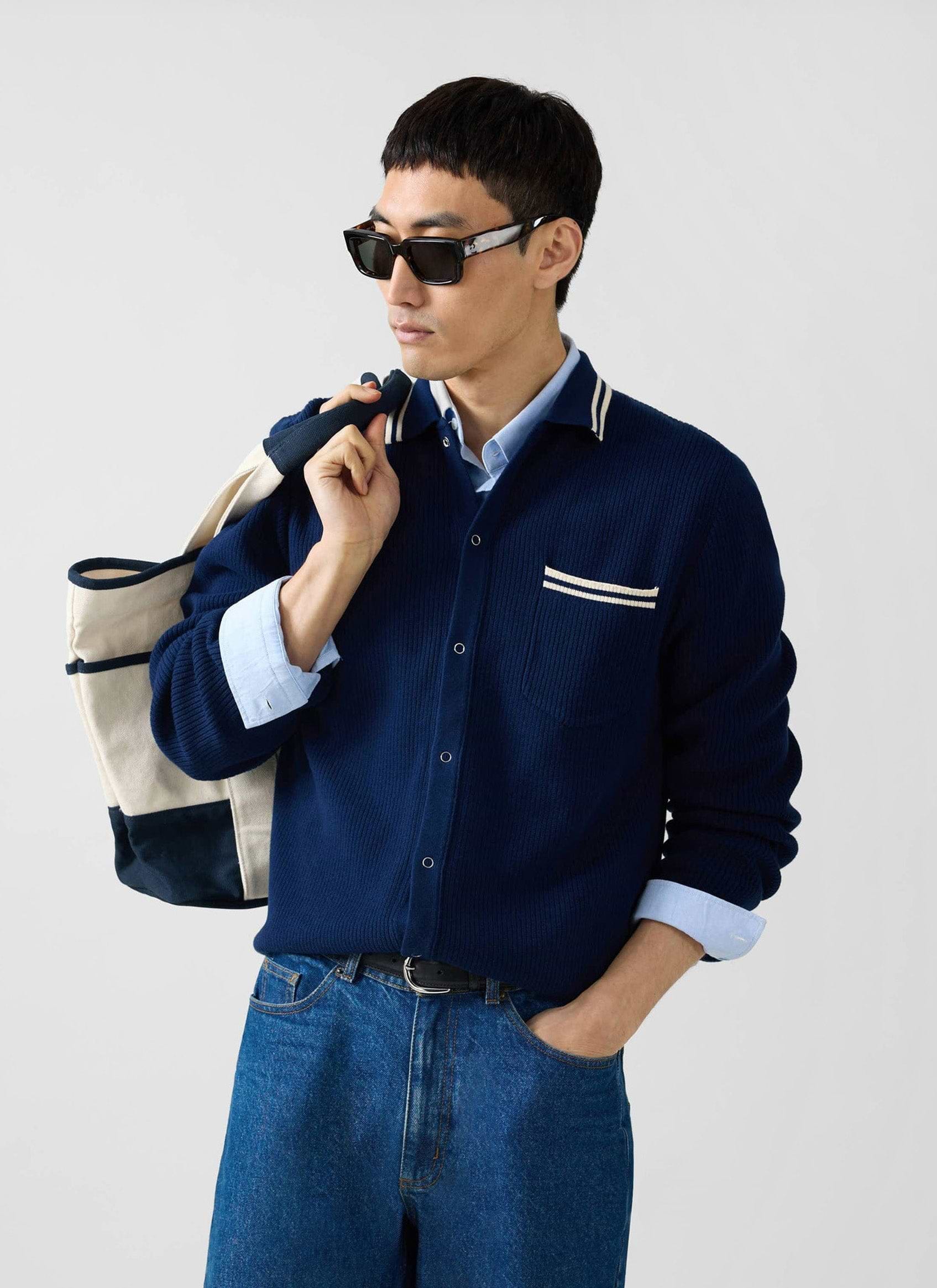 A man models the Percival Pablo Cotton Knitted Shirt in navy, layered over a shirt and paired with jeans and sunglasses.