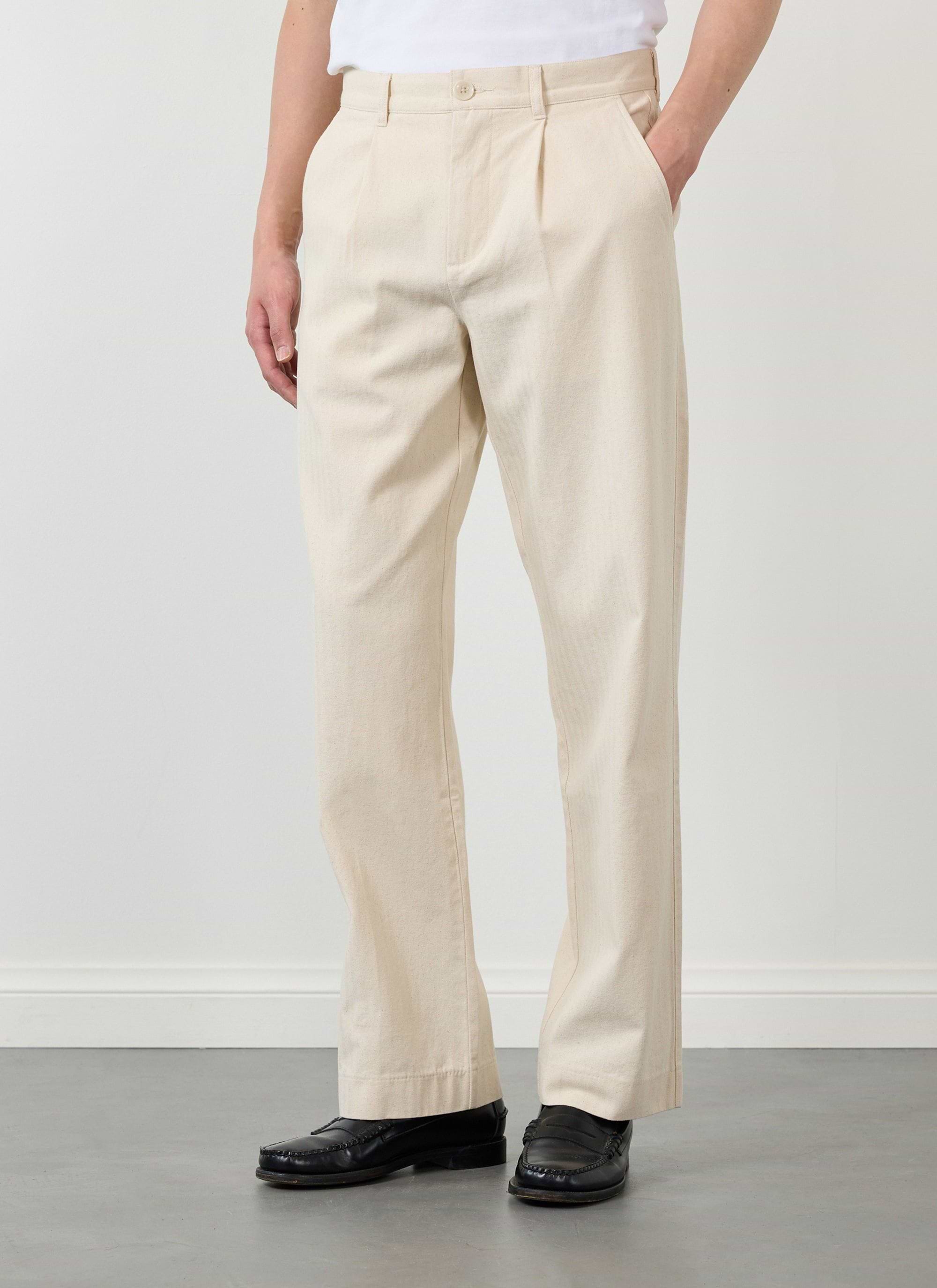 A model wears the Percival Program Pleated Trousers in ecru cotton with black loafers.