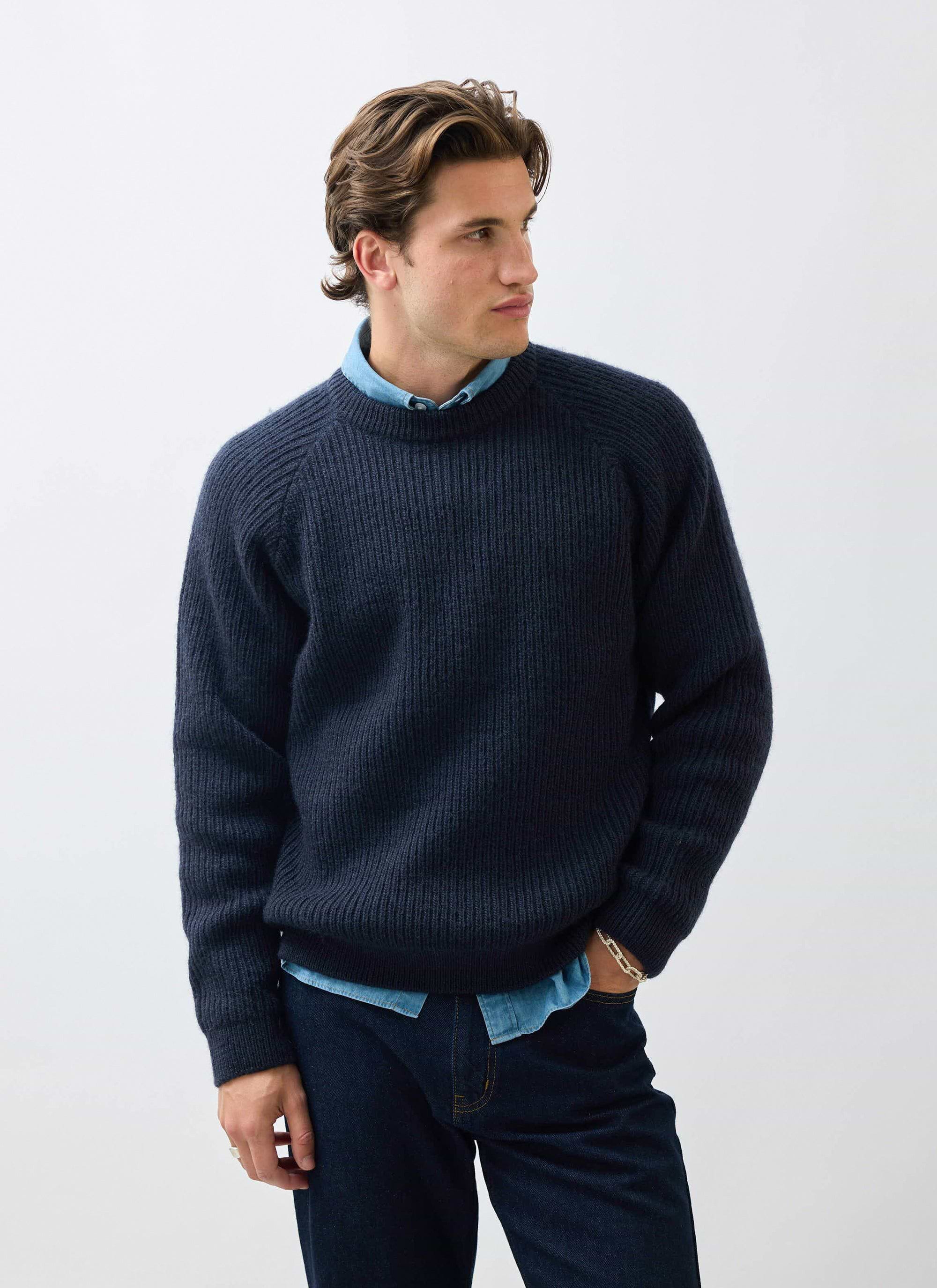 A man models the Percival Fisherman Raglan Jumper in navy wool, layered over a denim shirt and paired with dark jeans.